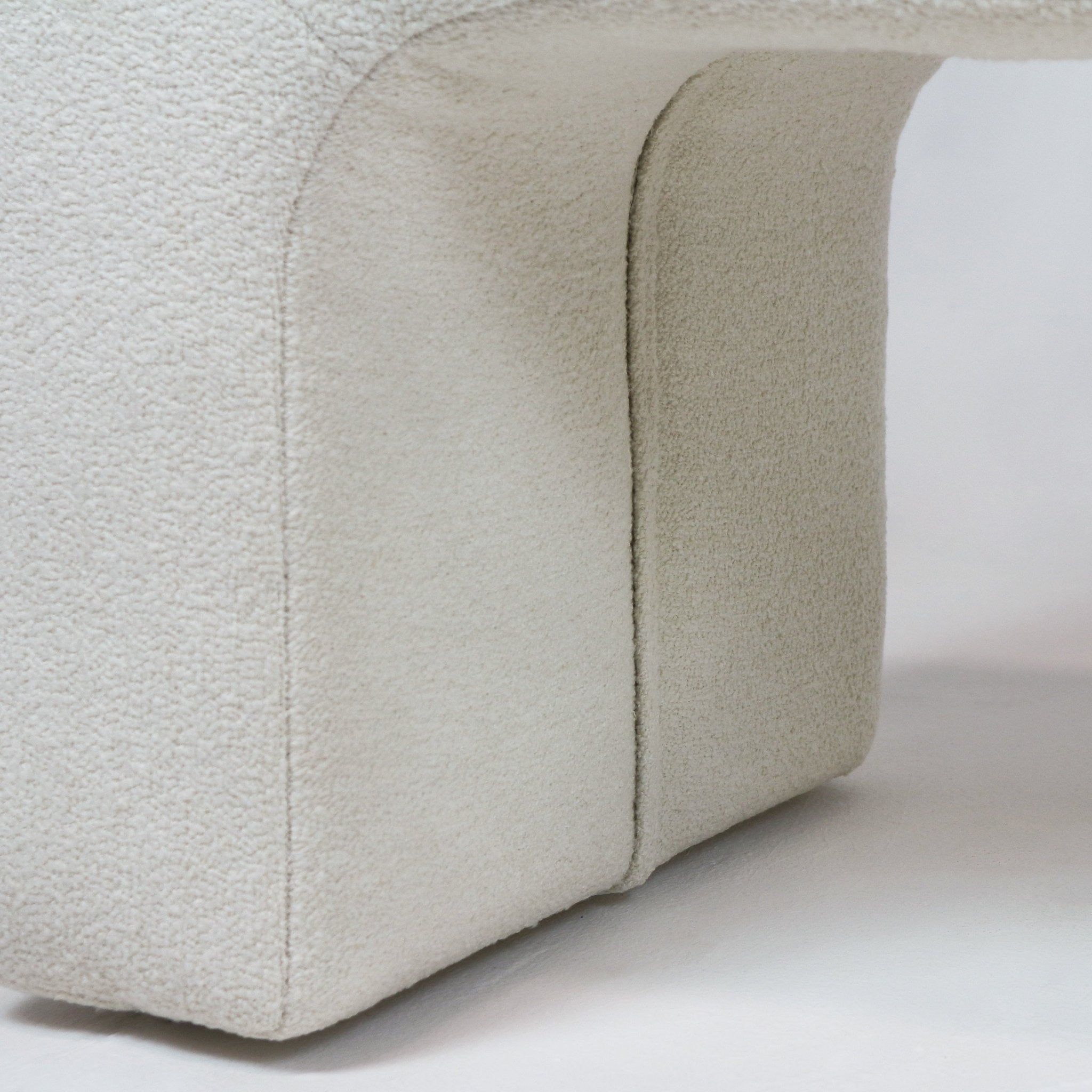 Close-up of cream boucle upholstery on the curved base of an end-of-bed bench, highlighting the textured looped fabric and a vertical seam where two upholstered panels meet.