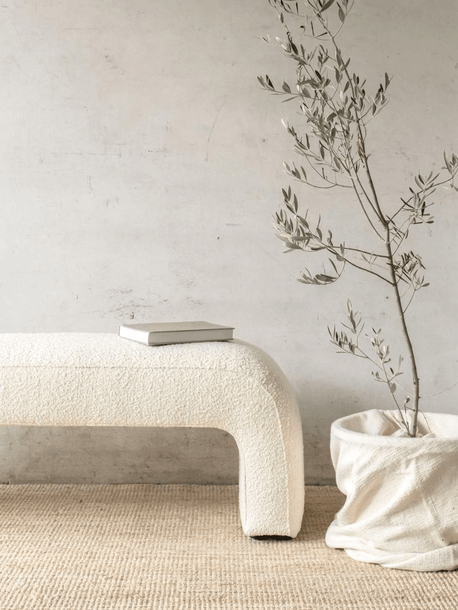 Cream boucle upholstered arched end-of-bed bench with a closed book on top, placed on a natural jute rug against a textured plaster wall, beside a potted olive branch in a soft linen-like fabric basket.