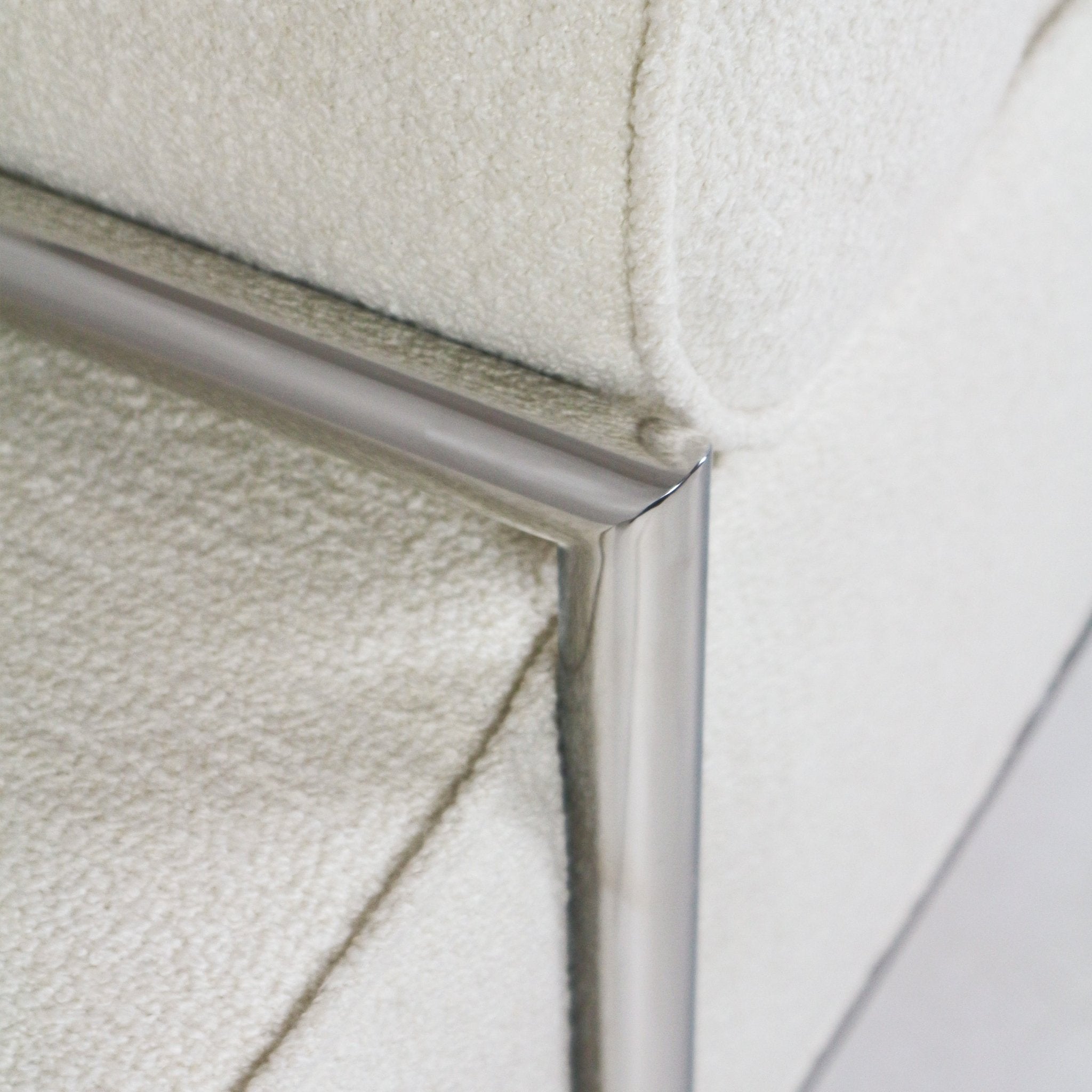Close-up detail of cream boucle upholstery and the polished stainless steel frame corner on a modern end-of-bed bench, showing fabric texture and metal finish.