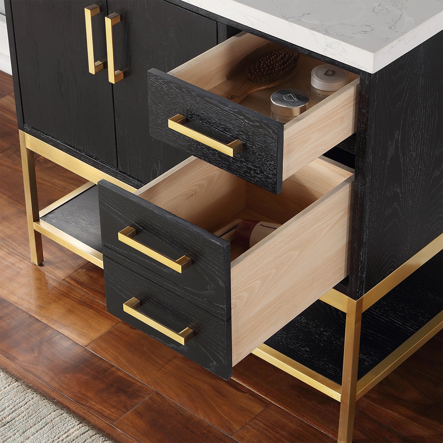 Close-up of a modern bathroom vanity with two open black-stained wood drawers showing light wood interiors, brass-tone rectangular handles and metal base, and a white stone countertop with toiletries inside the top drawer.