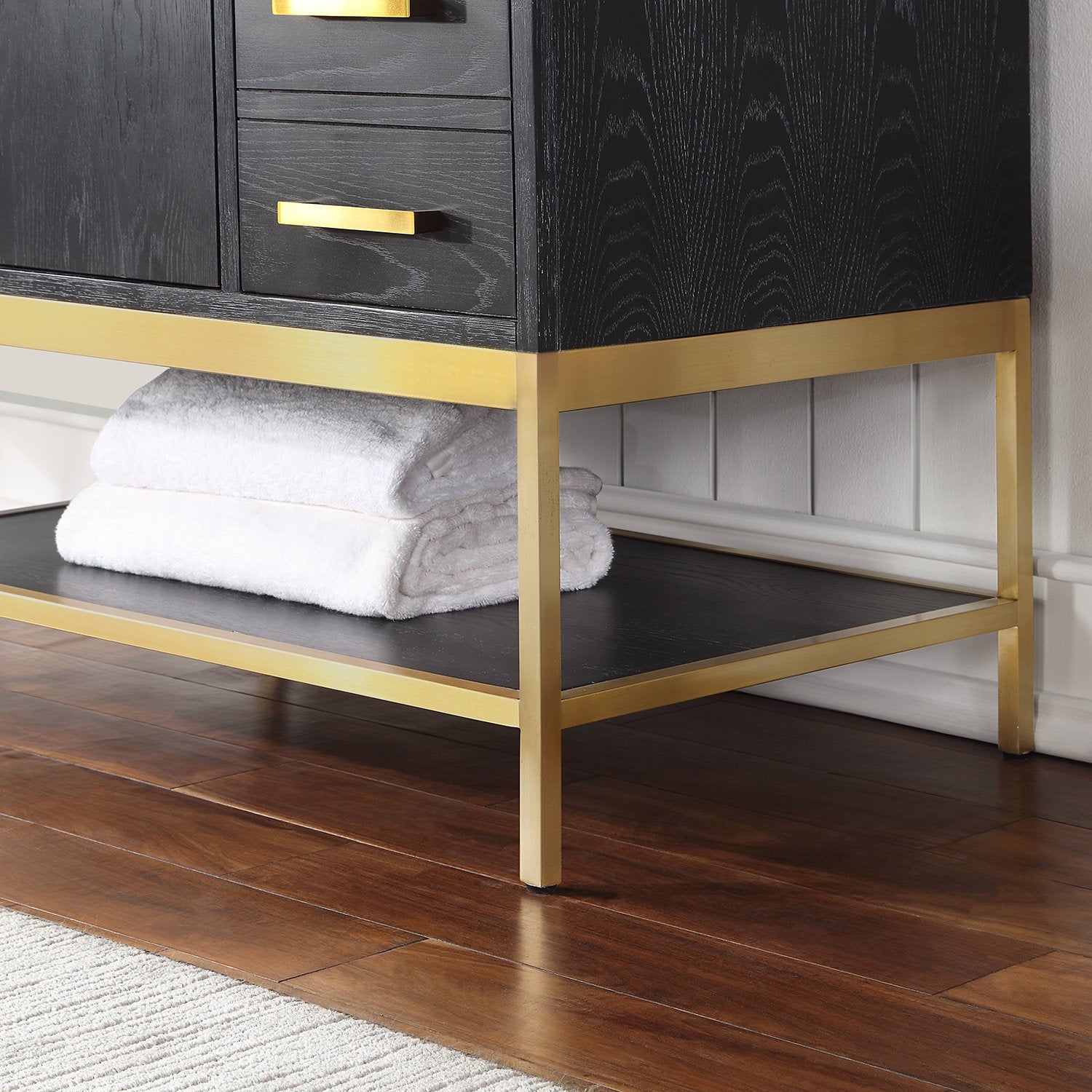 Close-up of a dark wood vanity lower section with a gold metal frame and legs, matching brass drawer pulls and an open shelf holding folded white towels on a wood floor.