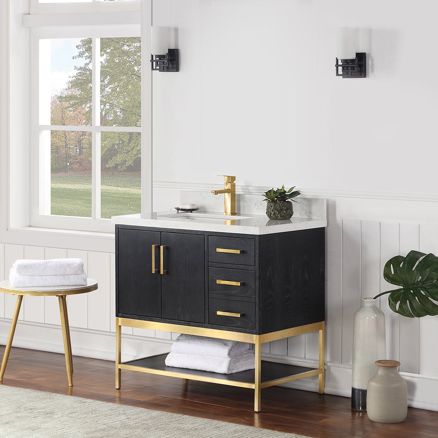 Modern black solid-wood vanity with white stone countertop and ceramic single sink, brass faucet and matching hardware, brass-framed open shelf with folded towels, styled with a potted plant and decorative vases against a white paneled wall.