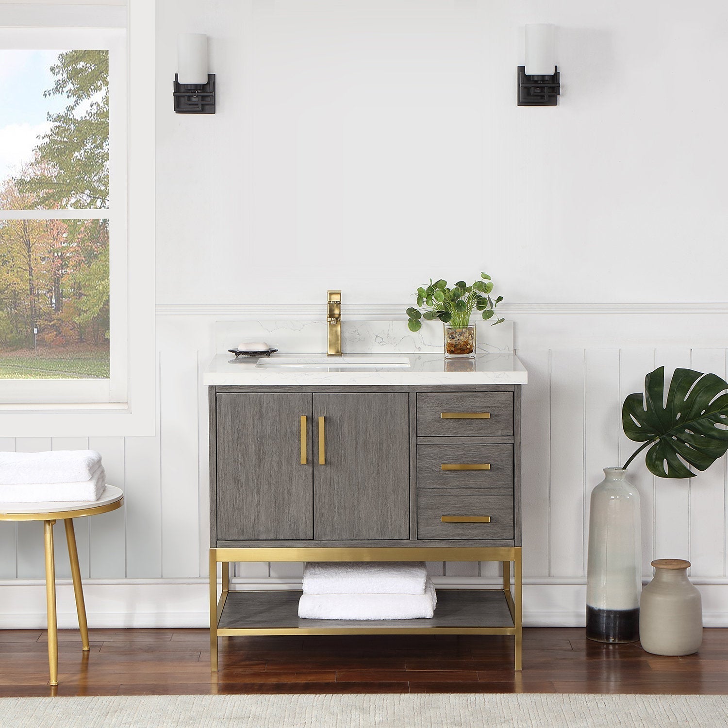 36-inch freestanding modern gray solid-wood vanity with stone countertop and white undermount sink, brass faucet, brass handles and base, two cabinet doors, three drawers and a lower shelf with folded white towels.