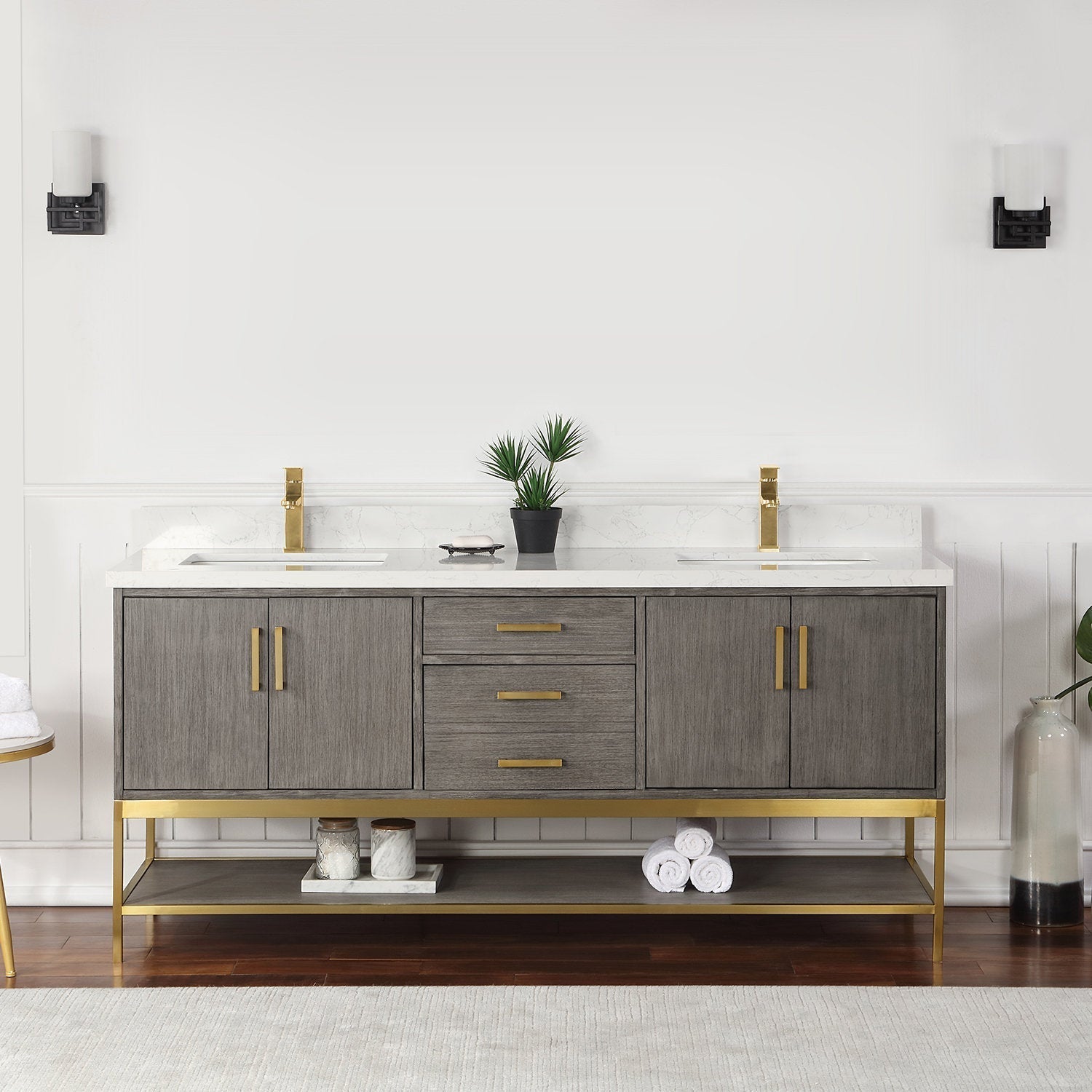 Solid wood 72-inch double-sink bathroom vanity in a stone finish with a white stone countertop, gold faucets and hardware, three center drawers, two cabinet doors and an open lower shelf with rolled towels and jars.