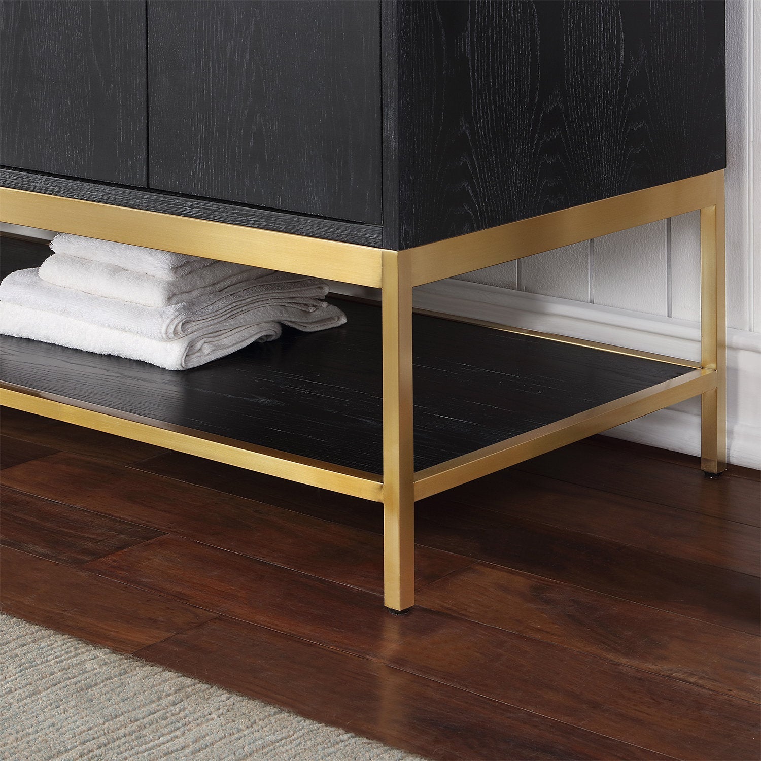 Close-up of the lower corner of a modern solid wood bathroom vanity in stone finish, showing a gold metal base and open lower shelf with folded white towels on a dark hardwood floor by a white beadboard wall.