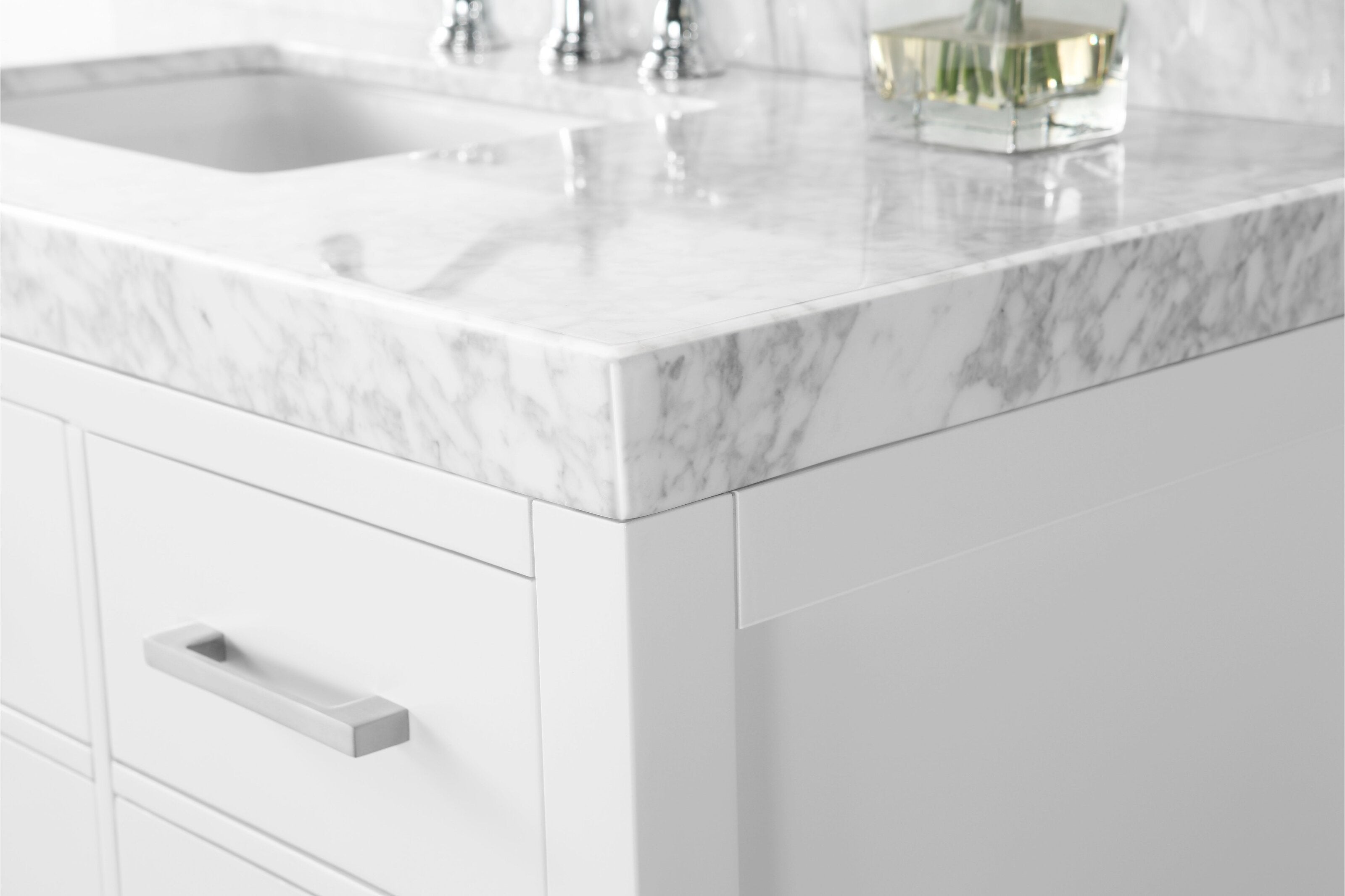 Close-up of a white solid wood bathroom vanity corner with a thick white marble stone top with gray veining, undermount sink and chrome faucet reflection, and a brushed metal rectangular drawer pull.