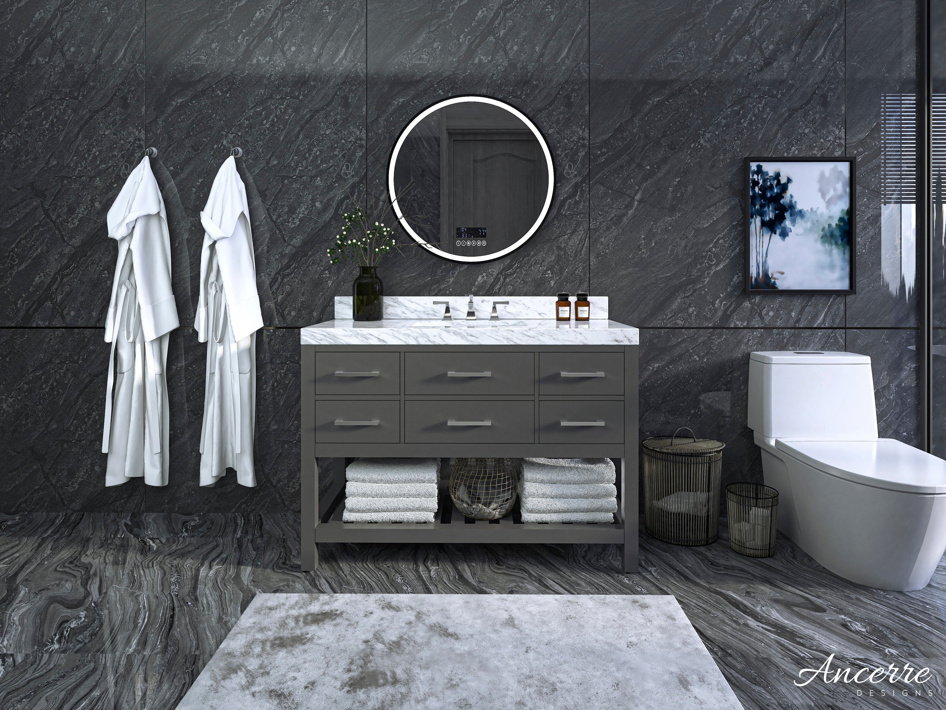 Modern 48-inch solid wood single-sink vanity with white marble stone top and stone-gray finish, round LED mirror above, folded towels on the open lower shelf, white robes on hooks and a white toilet against dark stone walls.