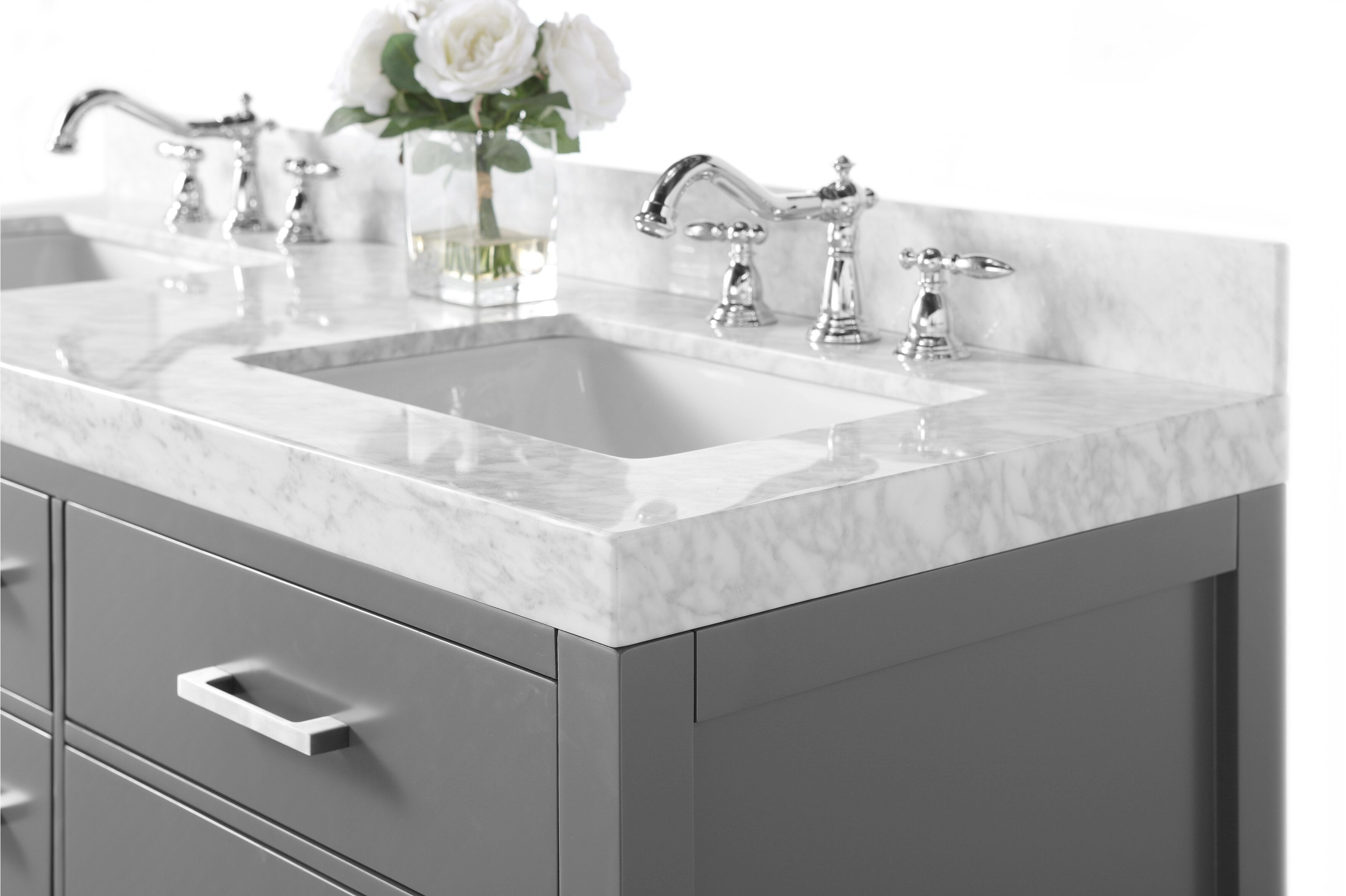 Close-up of a stone marble countertop with a rectangular undermount ceramic sink, polished chrome faucets and a stone-colored solid wood vanity with metal drawer pull and a glass vase of white roses.