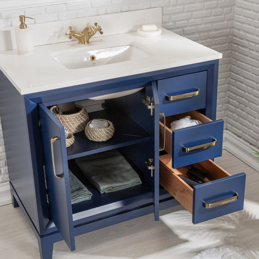 Modern navy-blue solid wood bathroom vanity with a white stone top and undermount ceramic sink, brass faucet and hardware; open cabinet and three drawers reveal woven baskets, folded towels and wood interiors.