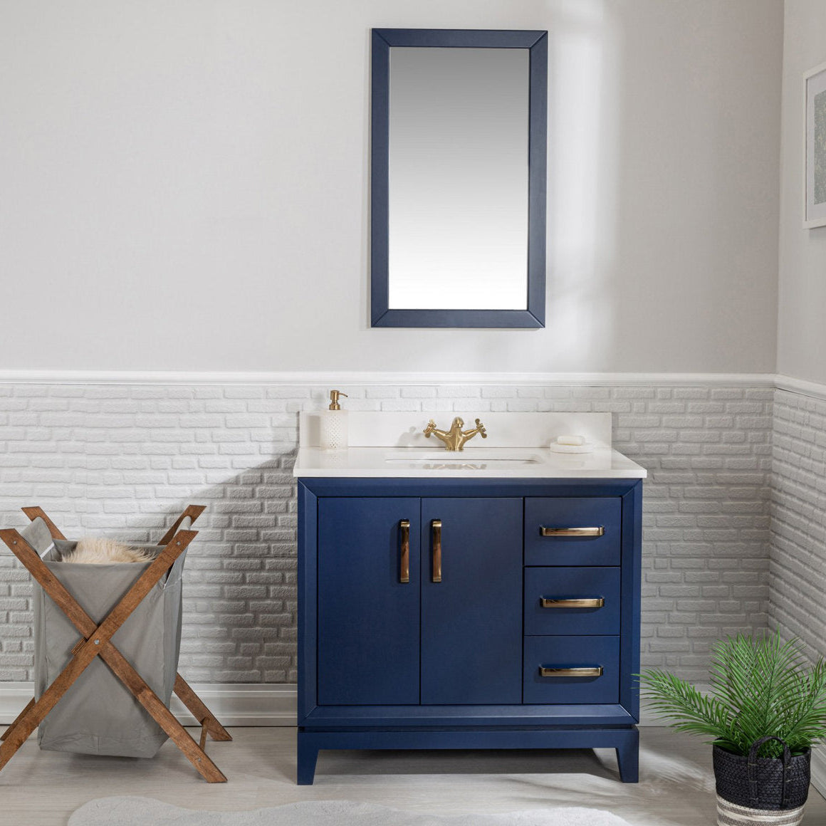 Freestanding navy blue solid wood bathroom vanity with white stone top and integrated sink, brass faucet and hardware, two-door cabinet with three drawers, matching navy framed mirror above, laundry hamper left and plant right.