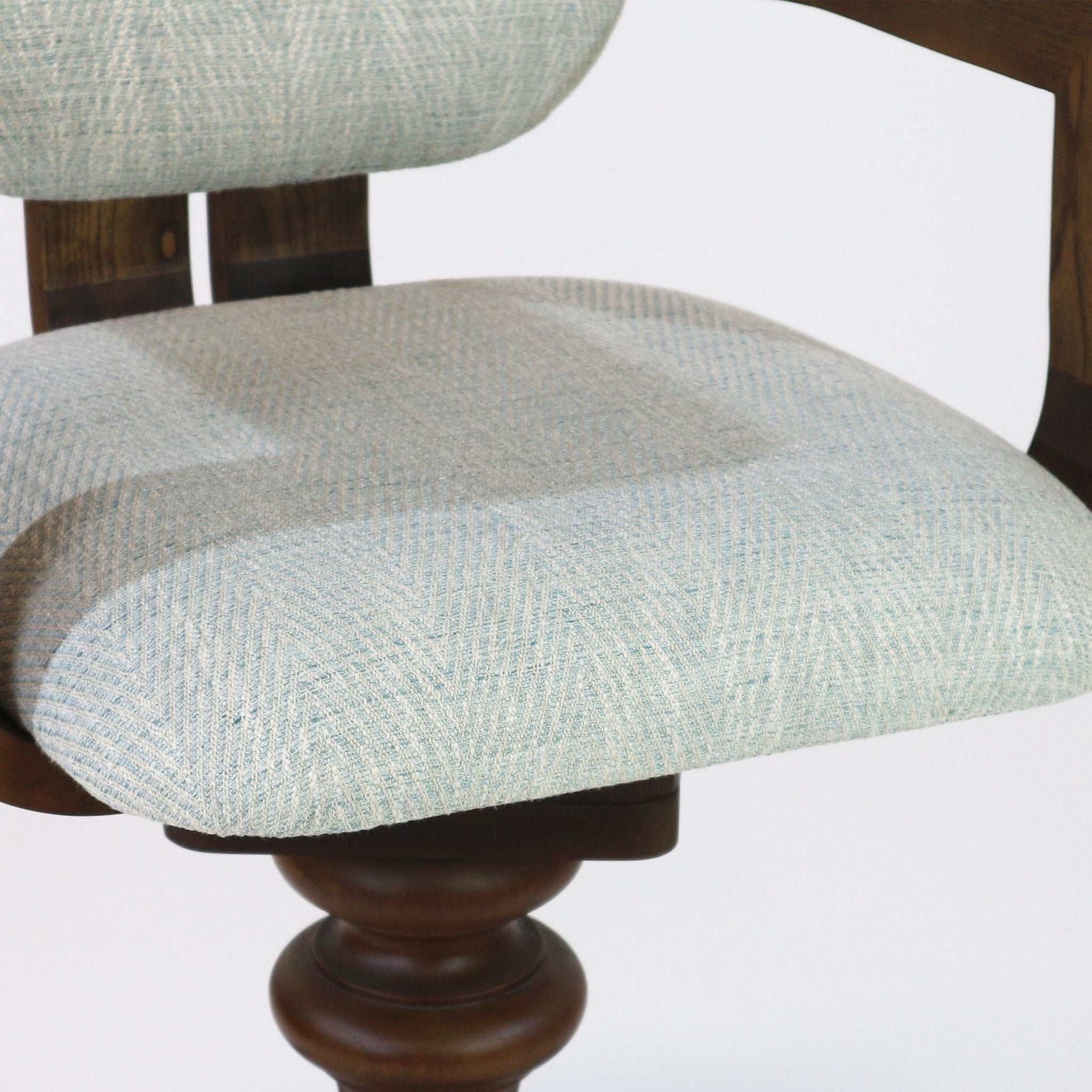 Close-up of a light blue-gray upholstered bar stool seat and partial back in textured woven fabric, showing cushioned tufting and a turned natural wood swivel base.