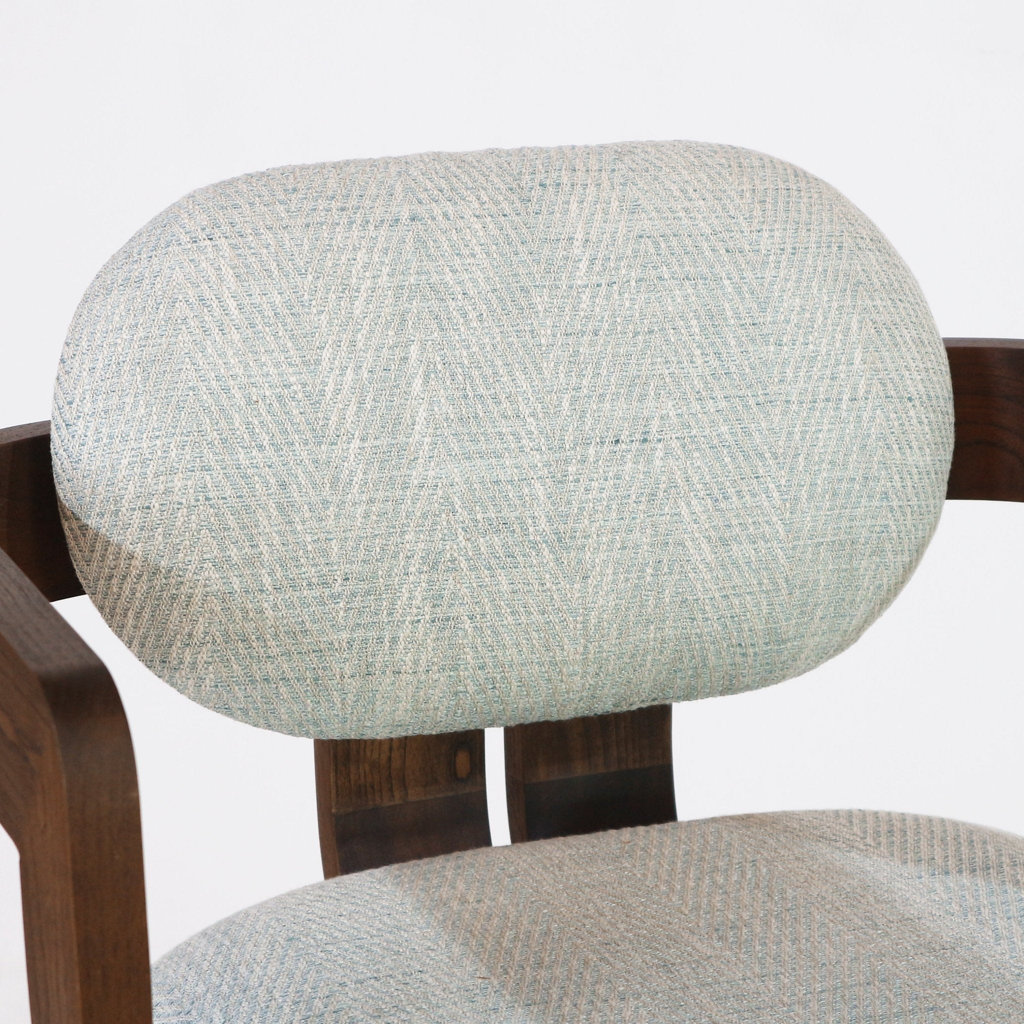 Close-up of a bar stool’s oval cushioned backrest and partial seat upholstered in light blue-gray woven herringbone fabric, mounted on a curved natural wood frame showing wood grain and fabric texture.