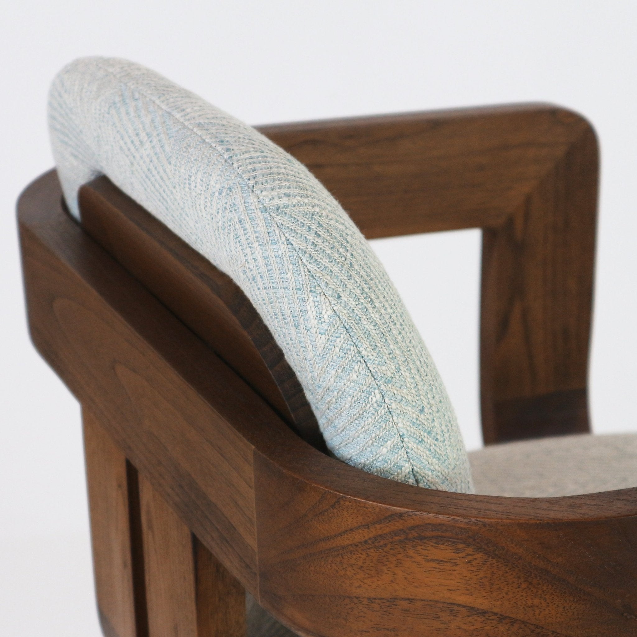 Close-up of a modern bar stool showing a light blue-gray herringbone fabric padded back resting against a curved natural walnut wood frame and arm, highlighting the upholstery texture and warm wood grain.