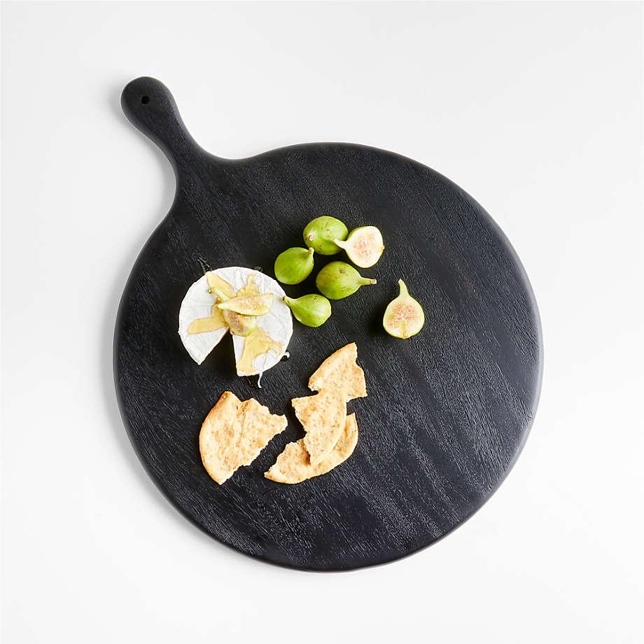 Top-down view of a round black teak butter board with a handle holding a wheel of brie cheese drizzled with honey, halved green figs and broken crackers on a white background.