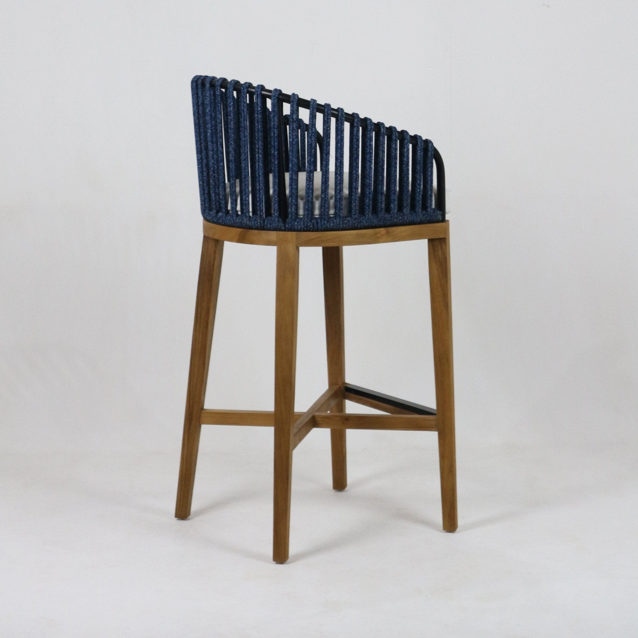 Side view of a teak-framed outdoor bar stool with a blue woven rope back, grey seat cushion, tapered natural wood legs and a black metal footrest.