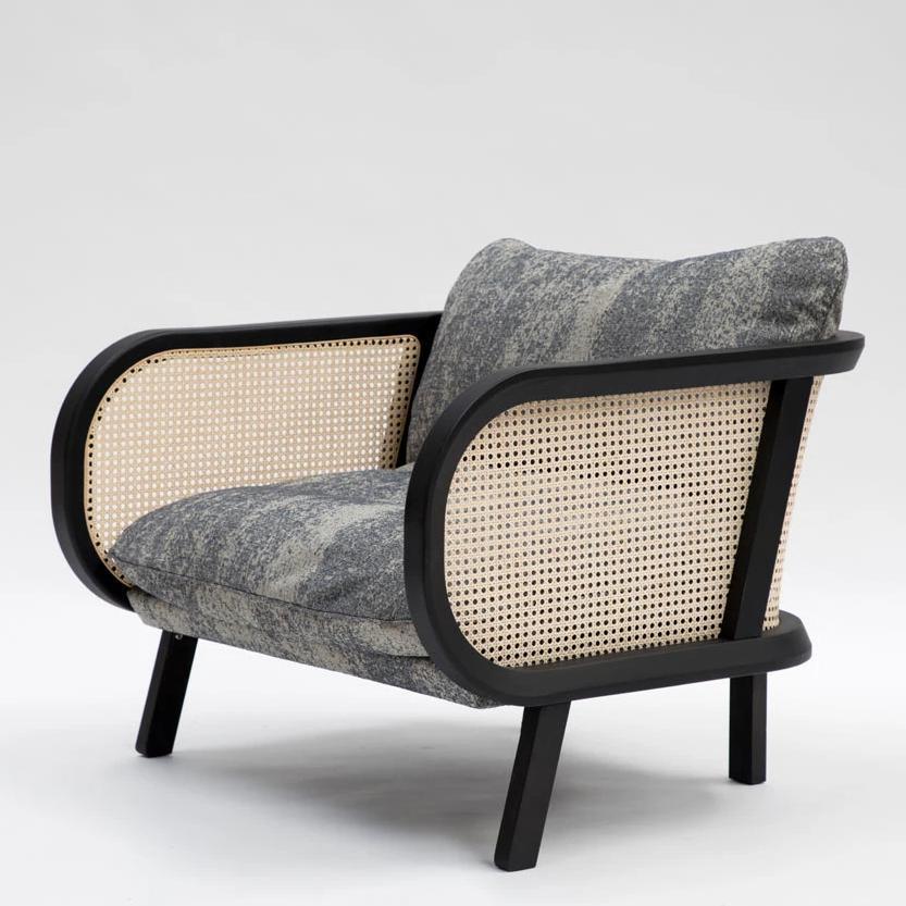 Contemporary lounge chair with a black wooden frame and natural woven cane side panels, upholstered in textured gray fabric cushions, featuring rounded curved arms and angled legs against a white background.
