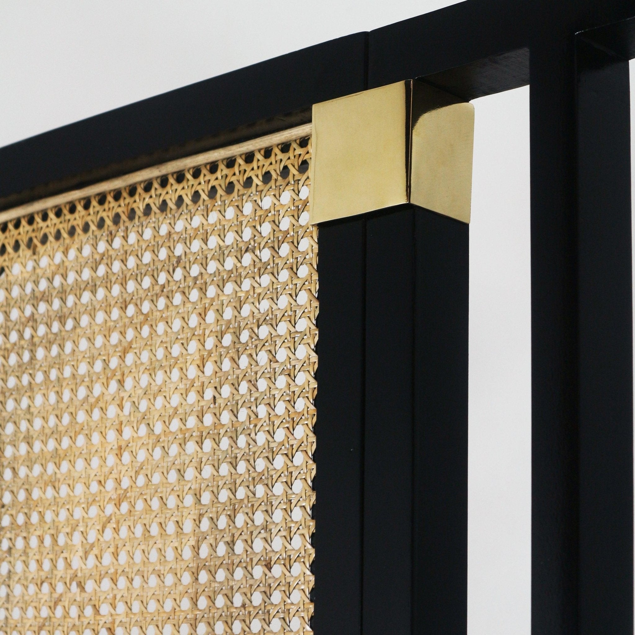 Close-up of a black framed headboard corner showing natural rattan cane weave panel and a polished brass corner cap on the black frame.