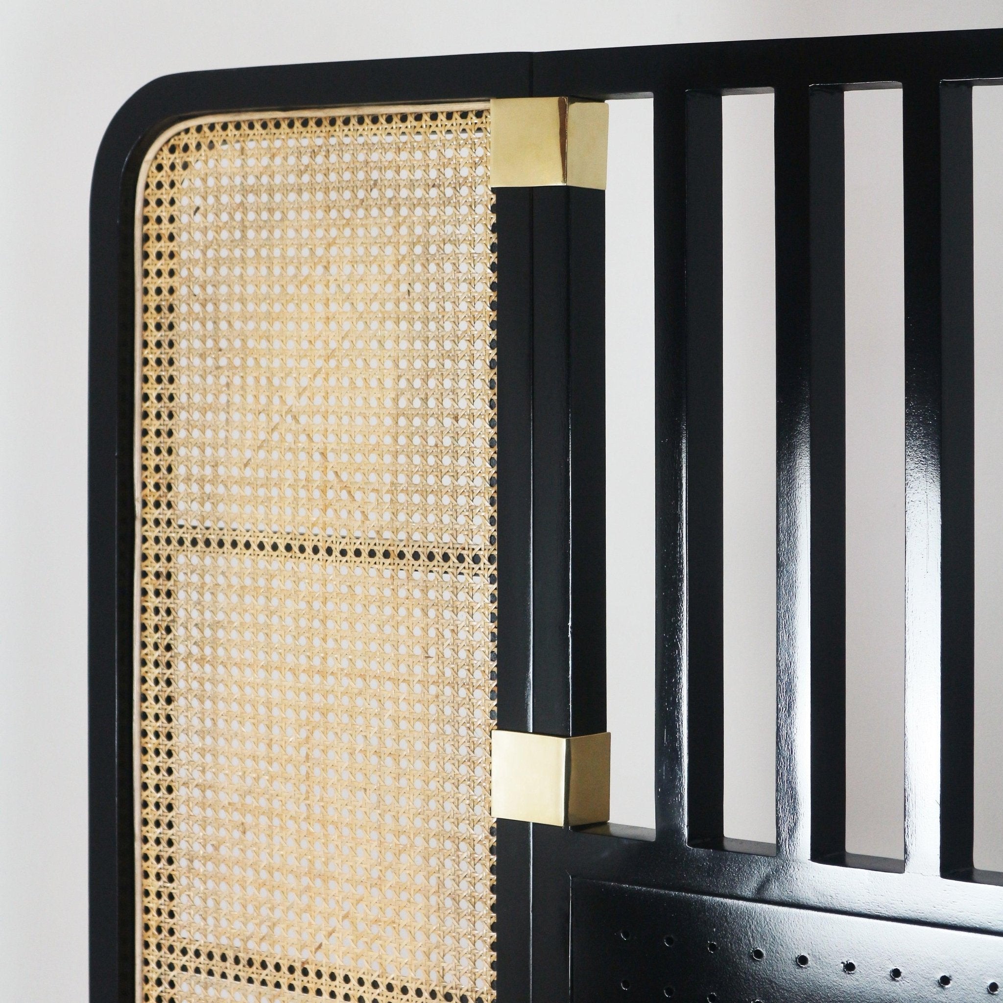 Close-up of a boho oak and rattan king headboard with a black painted frame, natural woven cane panel, vertical slats and polished brass corner accents showing the cane weave and joinery detail.