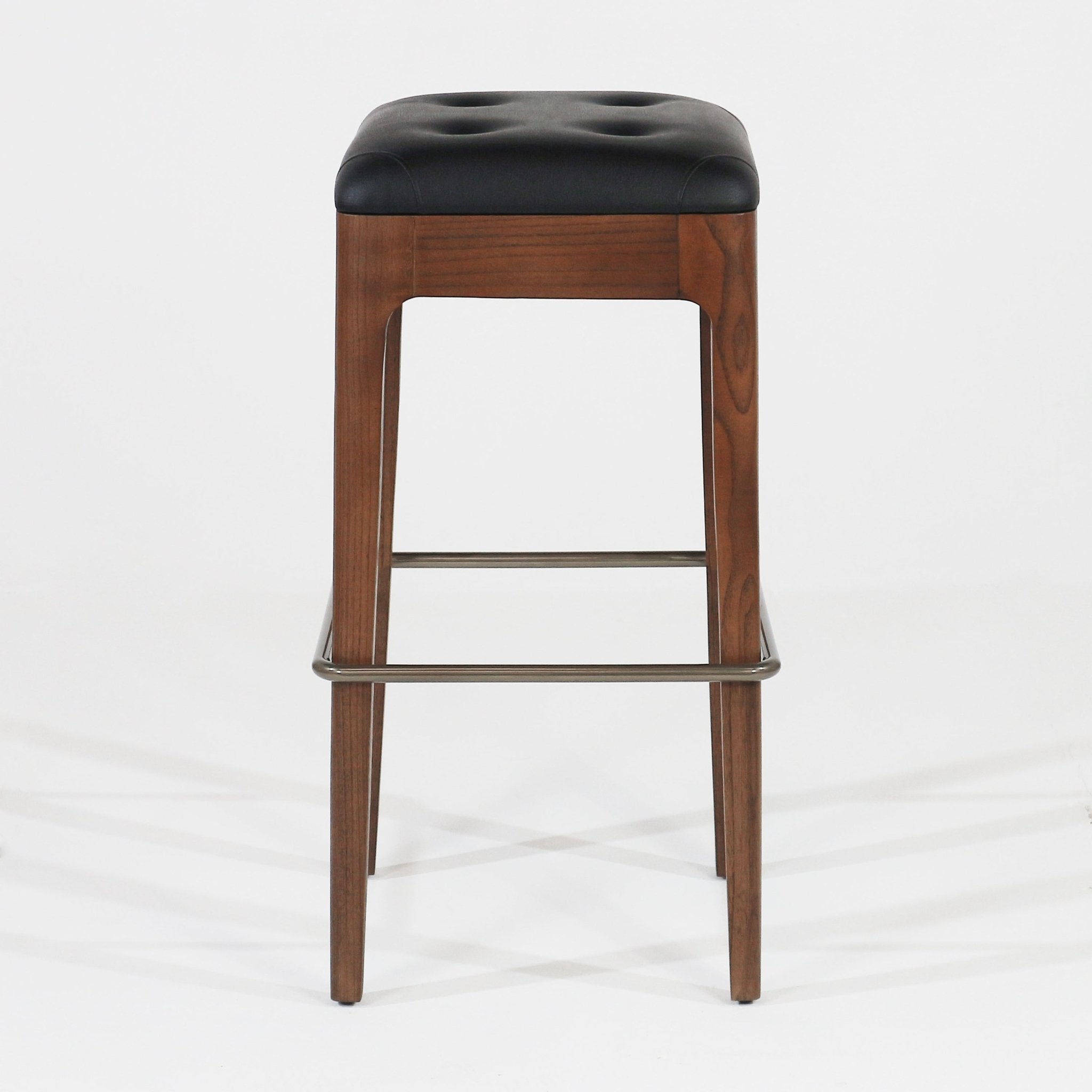 Front view of a modern backless stool with a tufted black leather seat, walnut wood frame and tapered legs, and a dark metal footrest.