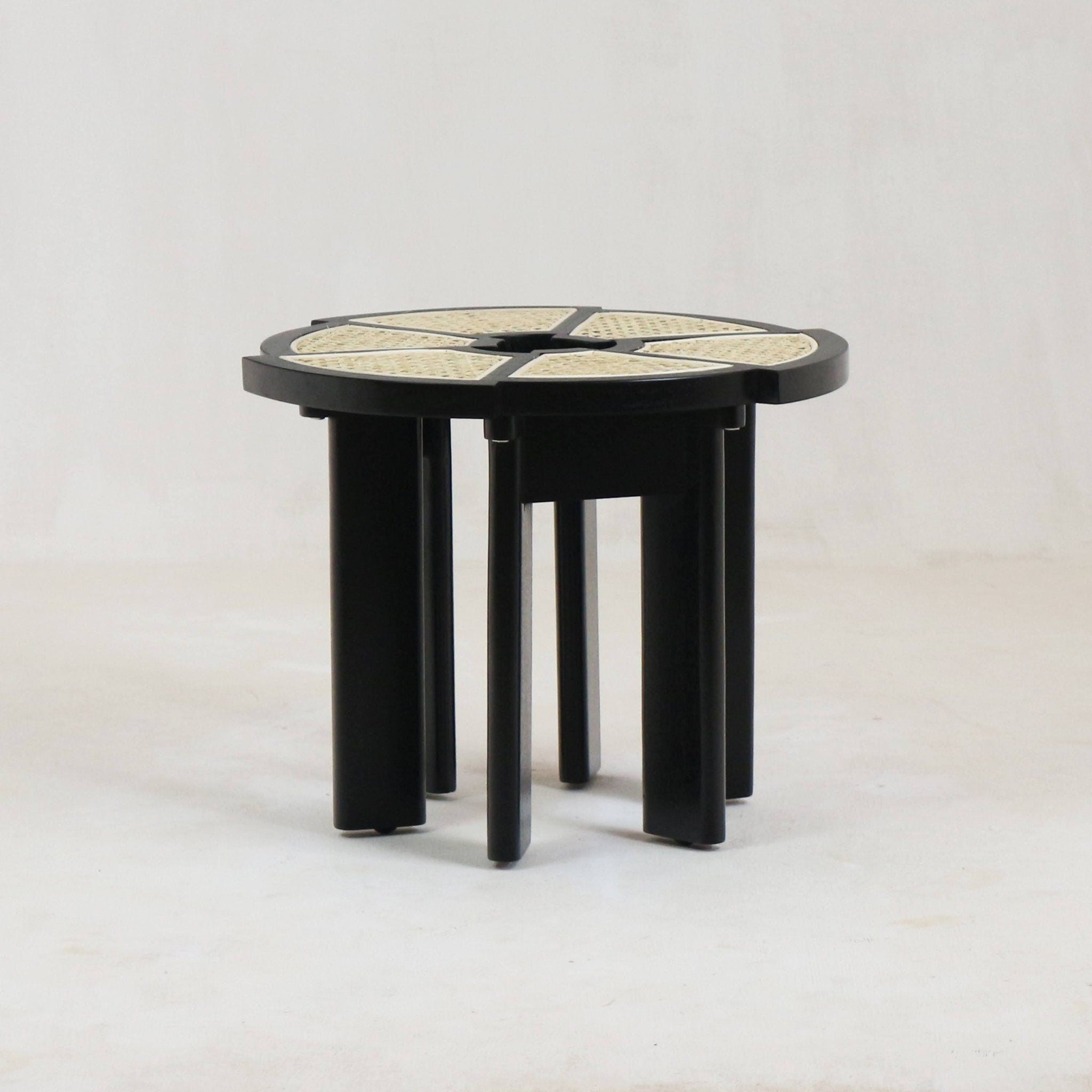 Round modern side table with a black gloss wooden frame and segmented natural cane top, supported by clustered black cylindrical legs.