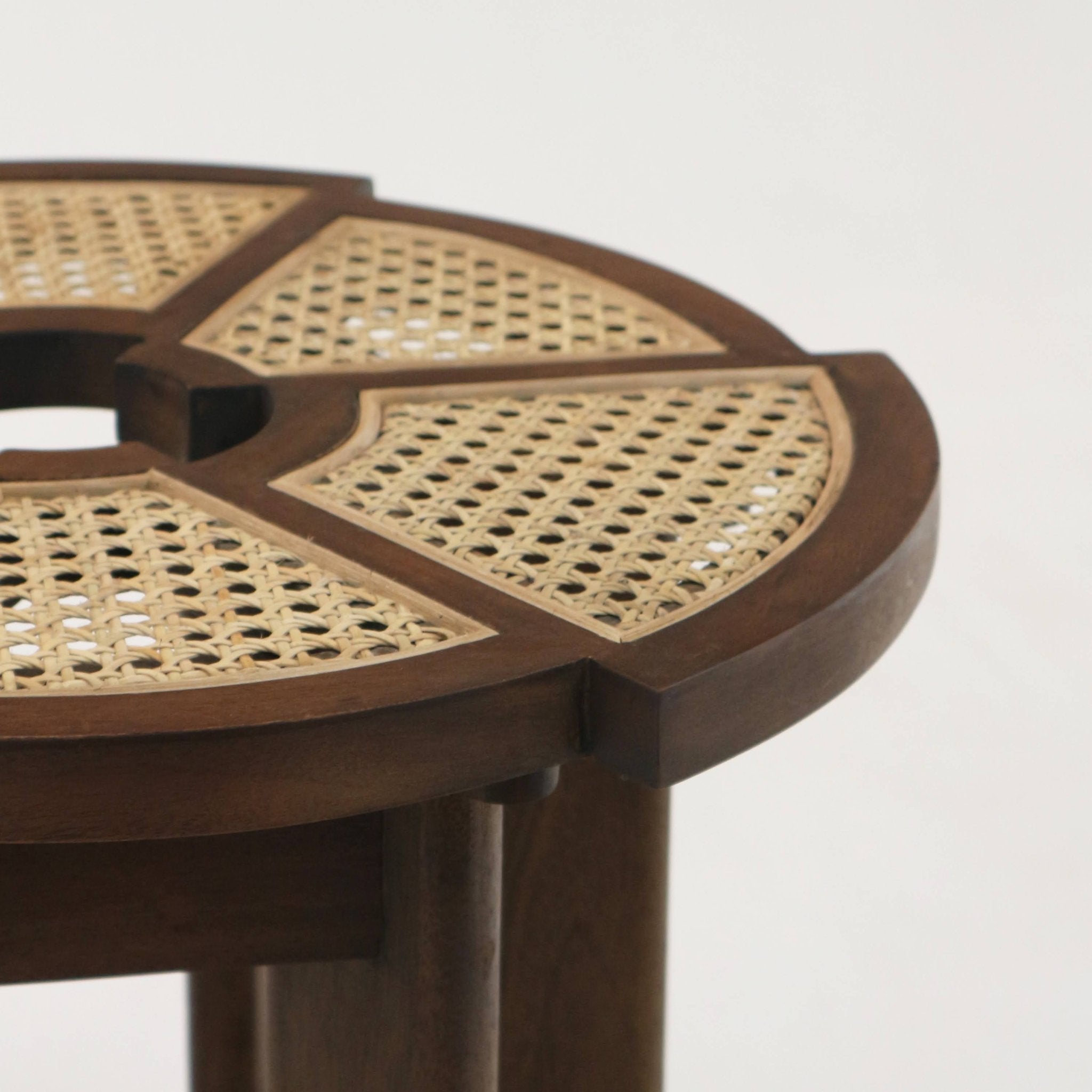Close-up of a round teak-brown side table top with woven natural cane inserts set into a solid walnut-toned wood frame, showing segmented cane panels, rounded outer edge and a portion of the cylindrical leg beneath.