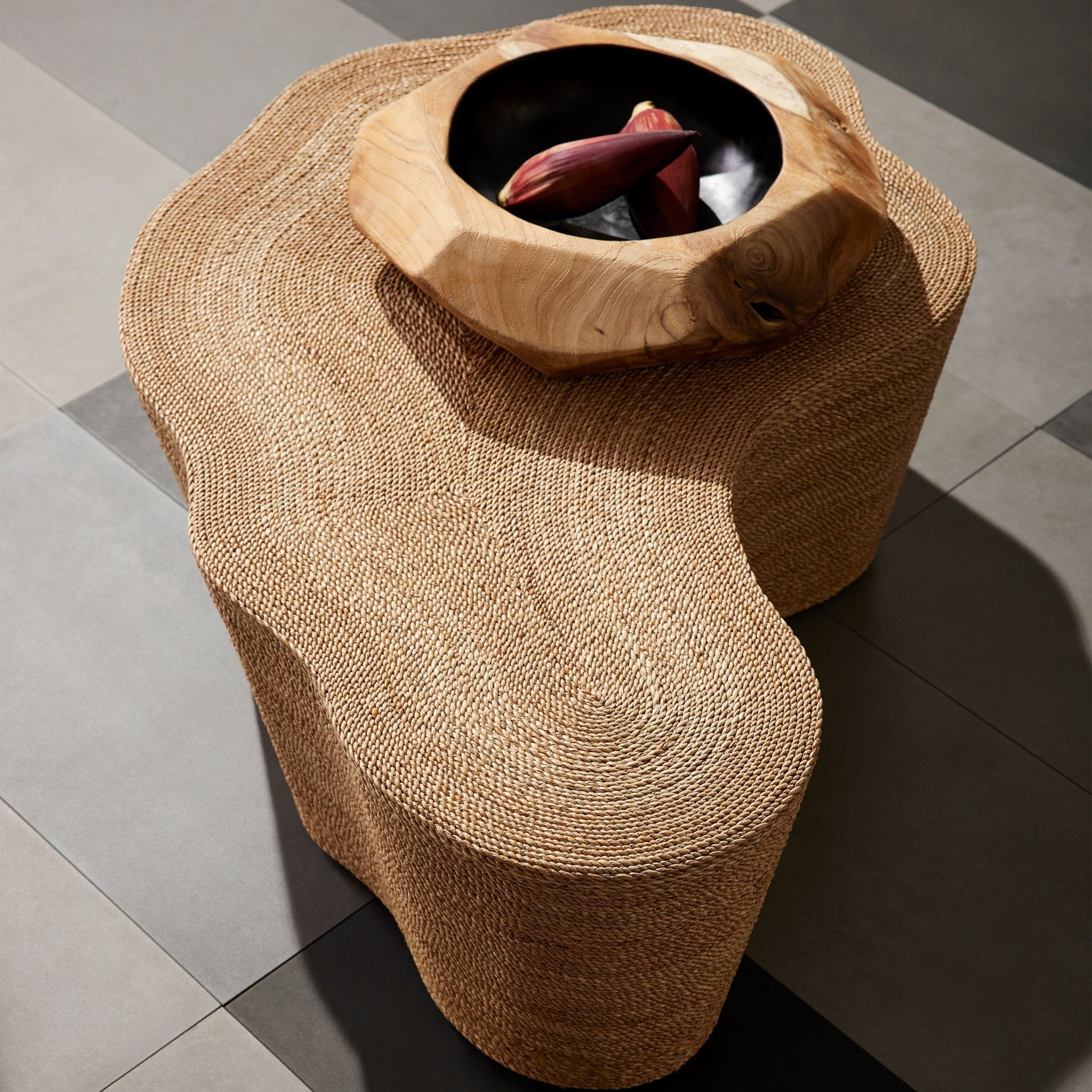 Top-down view of a natural woven rope coffee table with an undulating silhouette, topped by a carved wooden bowl with a black interior holding two purple tropical buds, set on gray tile flooring.