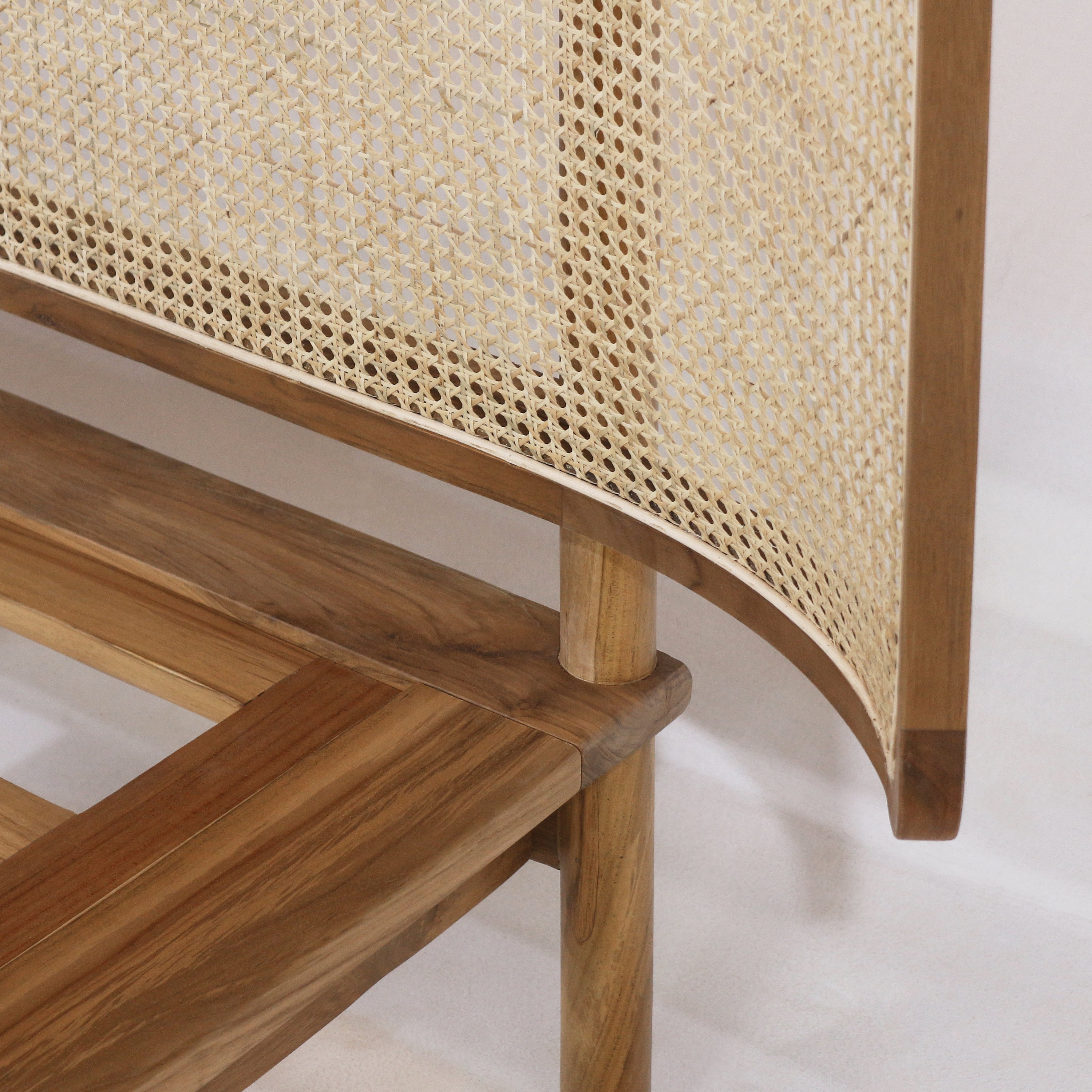 Close-up of a teak bed frame featuring a woven natural rattan cane headboard panel with a curved edge, slatted teak platform and rounded teak leg in a warm natural finish.