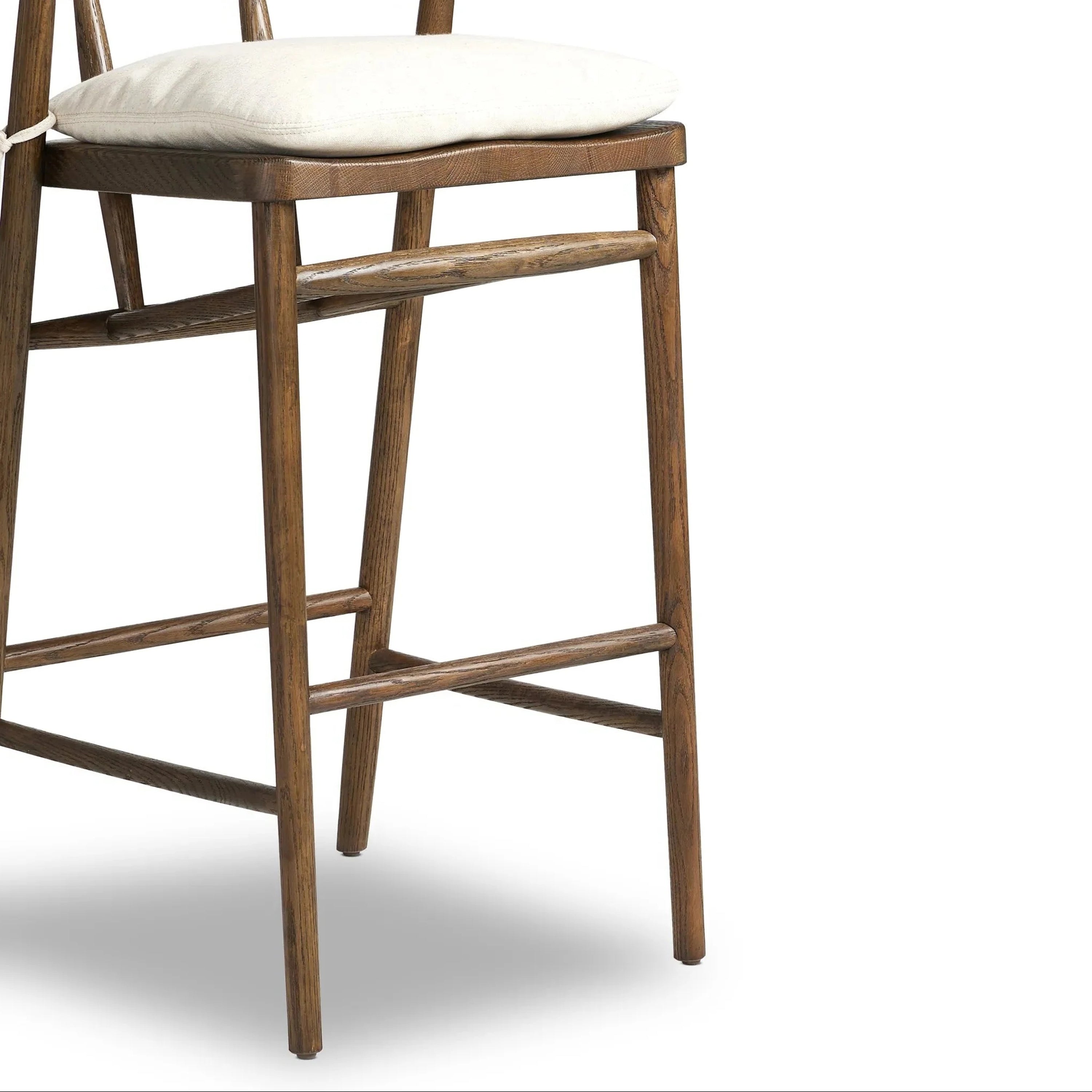 Close-up of a natural wood counter stool with a cream fabric cushion tied at the back, showing the shaped wooden seat, tapered legs and horizontal footrest in a rustic farmhouse finish.