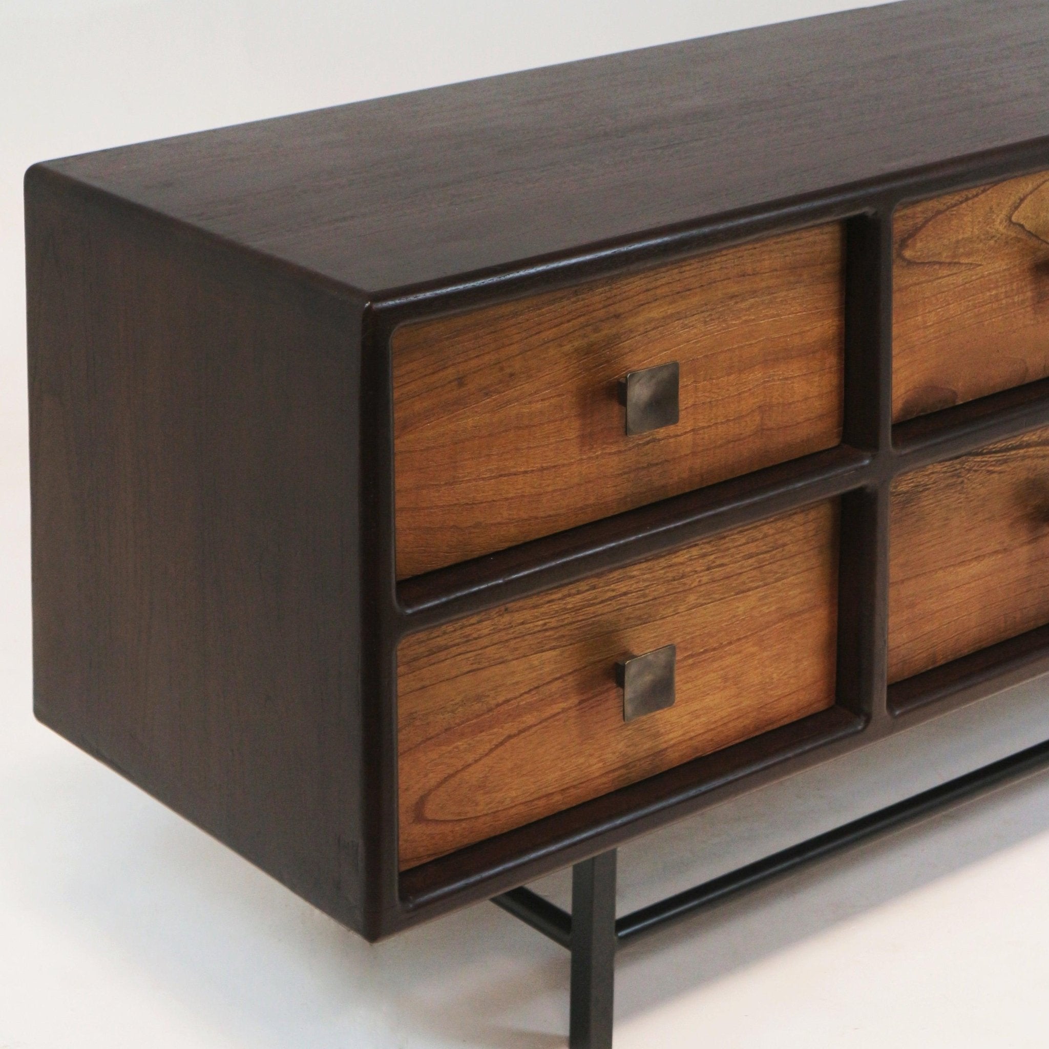 Close-up of a mid-century modern teak sideboard showing a dark-stained teak frame, warm teak drawer fronts with square iron knobs, recessed drawer openings and a slim black iron base.