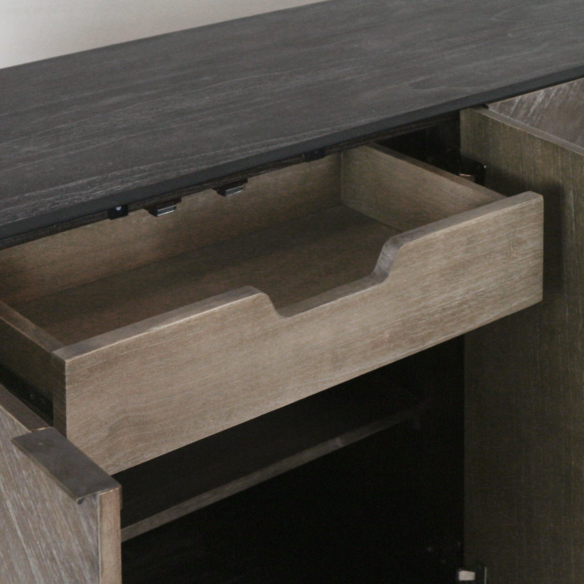 Close-up of an open teak sideboard drawer with a finger-notch cutout handle, showing the light teak interior, the black tabletop edge above, and a lower shelf inside the cabinet.