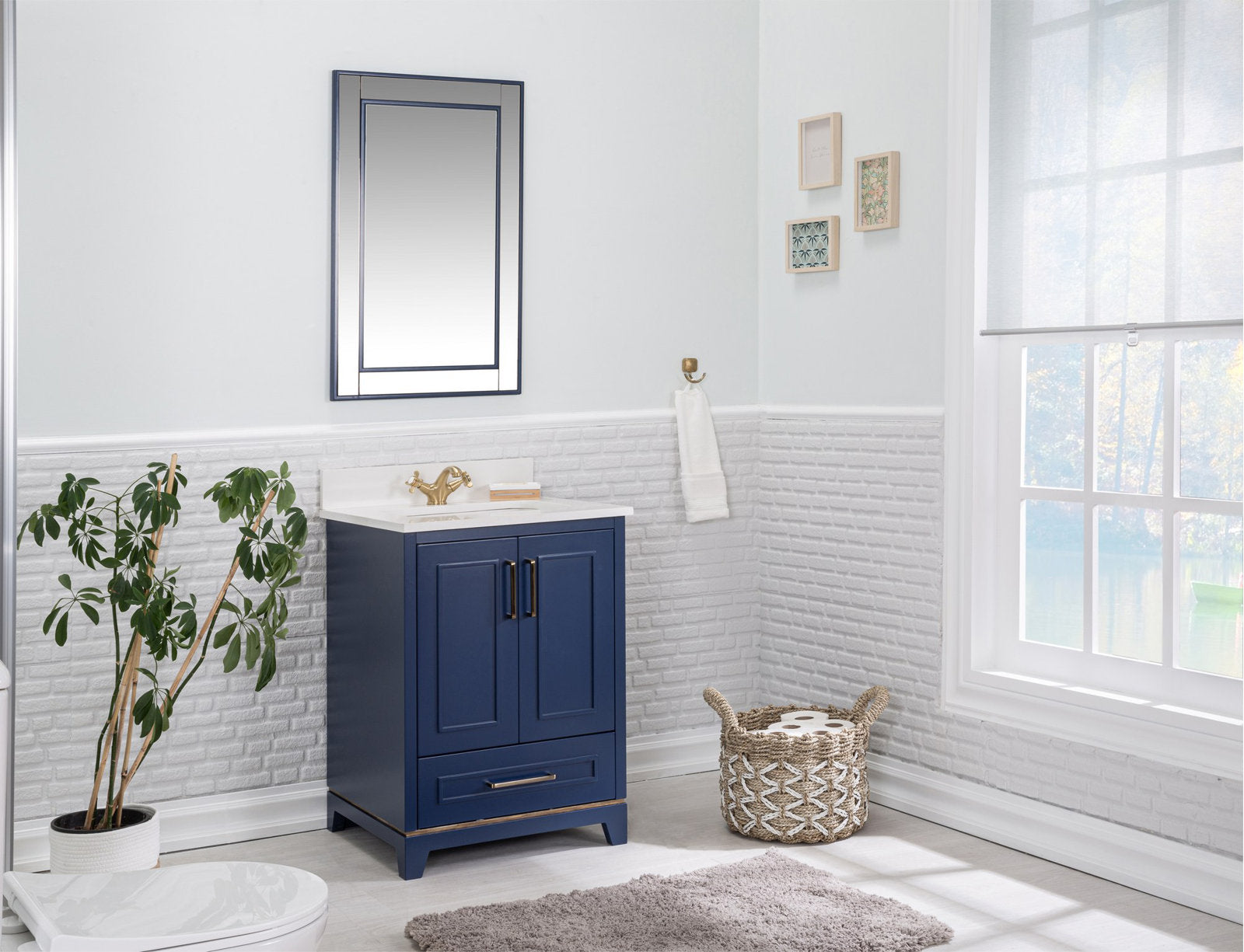 Freestanding modern solid wood single-sink vanity in navy blue with a stone countertop, brass faucet and hardware, two-door storage and drawer, styled with a mirror, towel hook, plant and woven basket in a bright bathroom.
