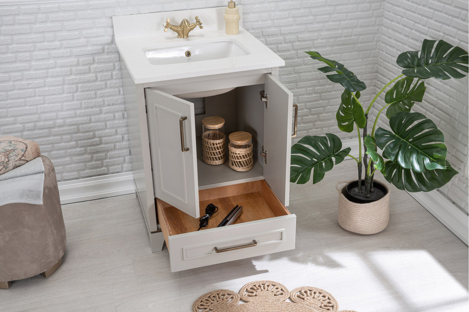 Modern stone-colored solid wood bathroom vanity with white stone top and ceramic sink, brass faucet and handles; cabinet doors open to show woven storage jars and a pulled wooden drawer holding a hair straightener.