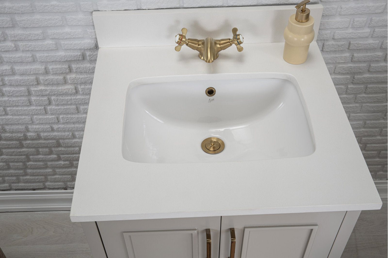 Top view of a modern stone-colored vanity showing a white ceramic basin set into a stone countertop with brushed brass faucet and drain, beige soap dispenser and a solid wood cabinet with brass handles against a textured white brick wall.