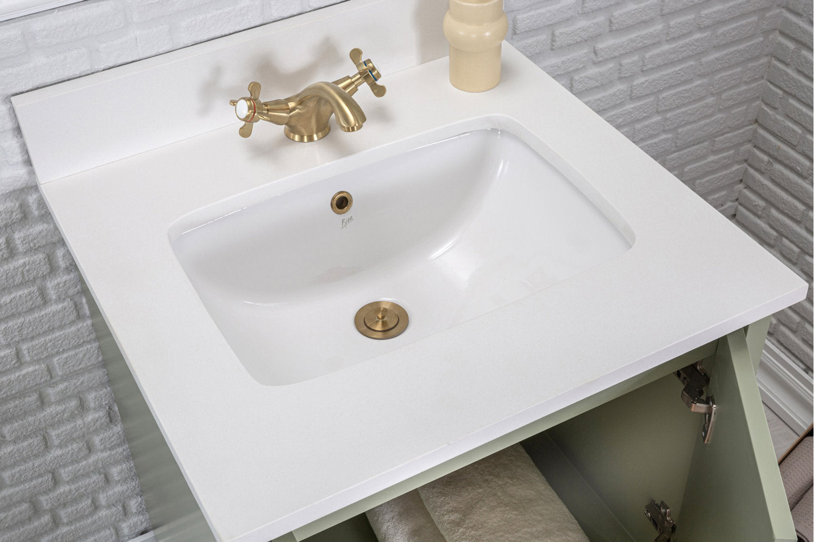 Overhead view of a modern bathroom vanity with a white stone countertop and recessed ceramic sink, brushed brass faucet and drain, and an open light-green solid wood cabinet showing a folded towel against a textured white brick backsplash.