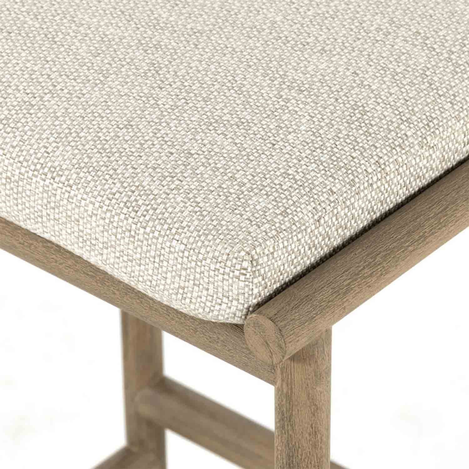 Close-up of a beige woven outdoor fabric seat cushion on a teak wood backless bar stool, showing the upholstered corner, teak frame joint and lower stretcher detail.