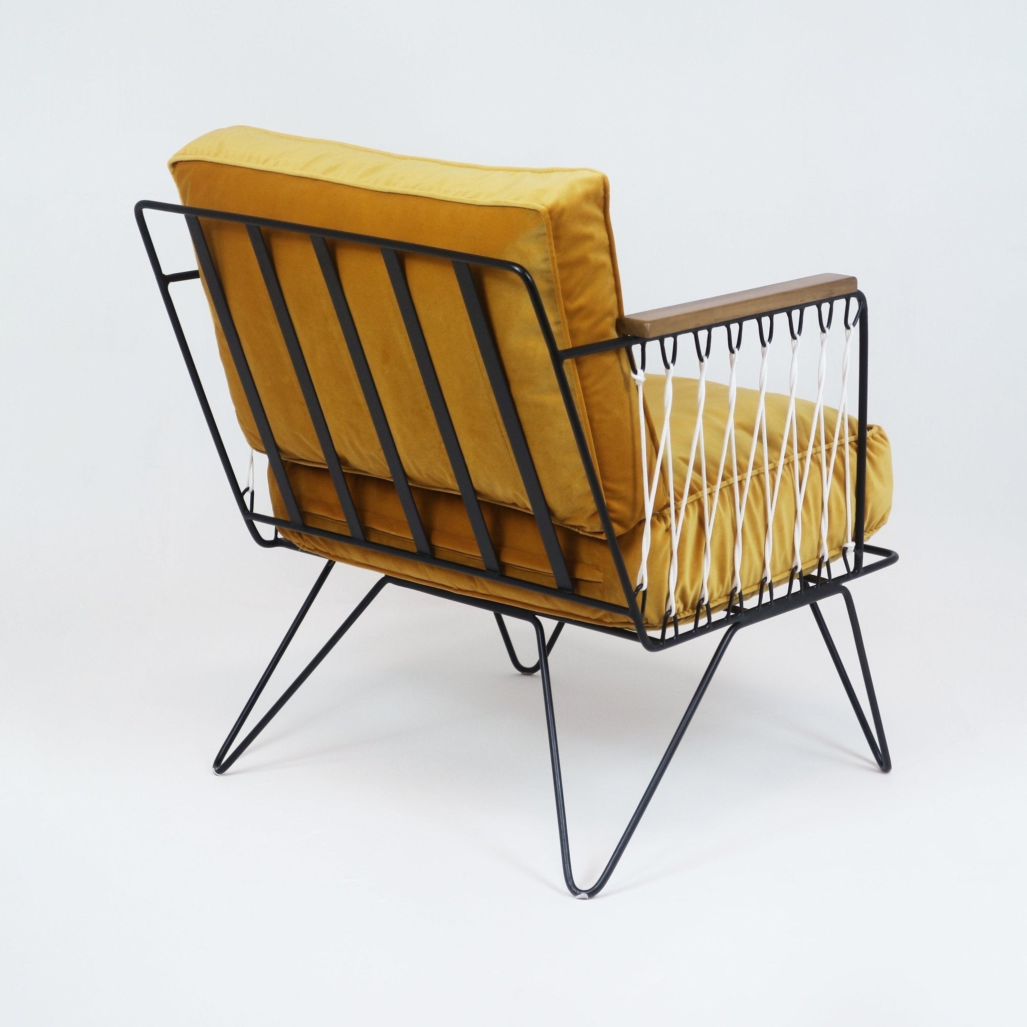 Three-quarter rear view of a modern lounge chair with mustard-yellow fabric seat and back cushions, black metal slatted frame, teak armrest and white rope side detailing mounted on hairpin metal legs.