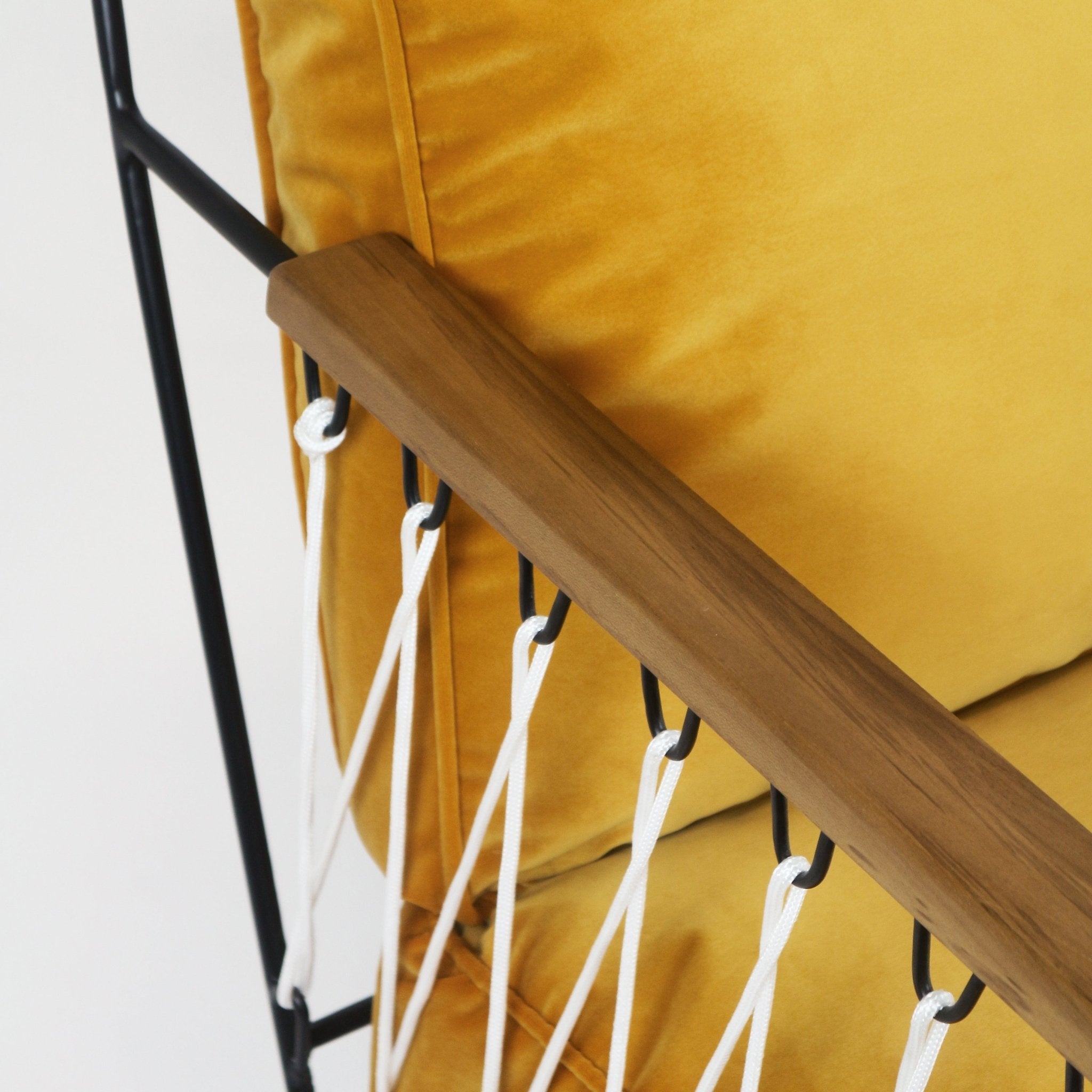 Close-up of a teak armrest on a black metal frame with white rope lacing beside a mustard yellow velvet cushion.