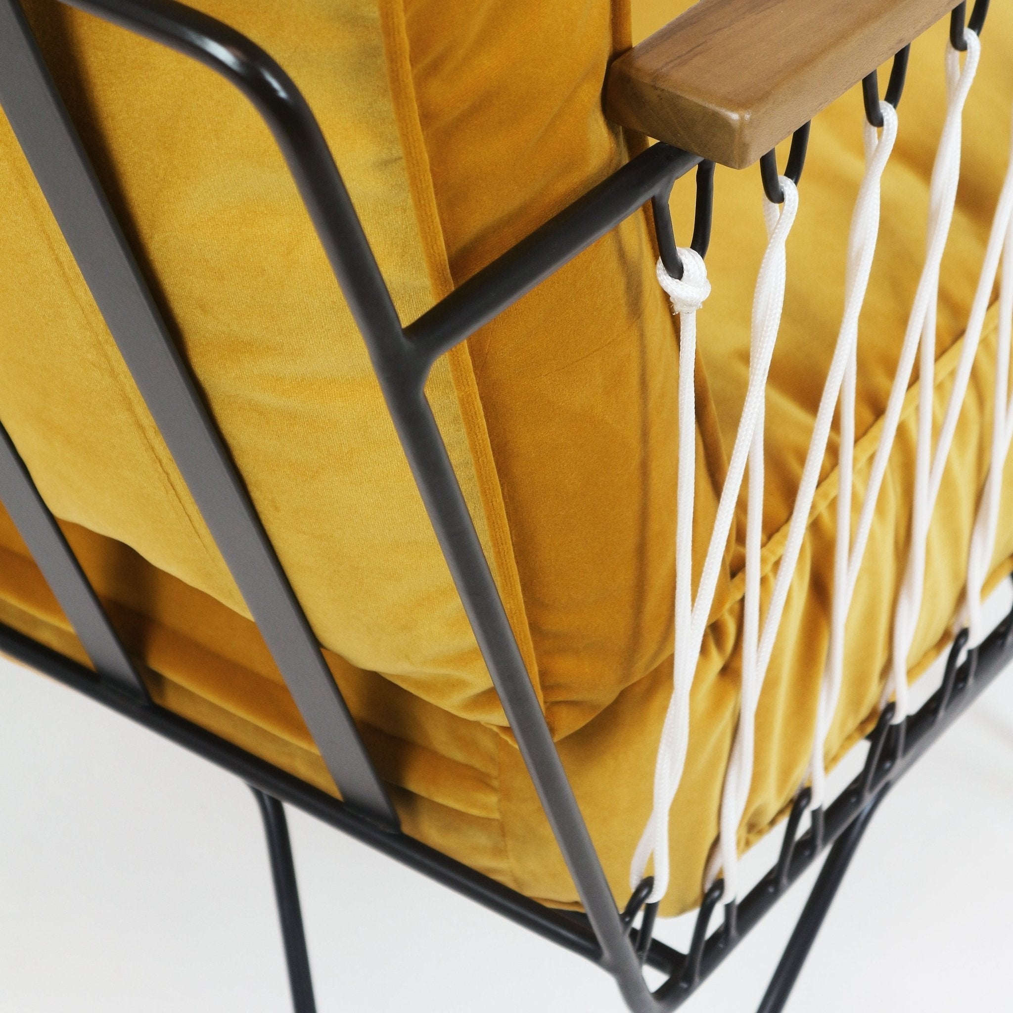 Close-up of a mustard yellow fabric cushion set in a black metal chair frame with white rope lacing and a teak armrest, highlighting upholstery piping and cord attachment details.