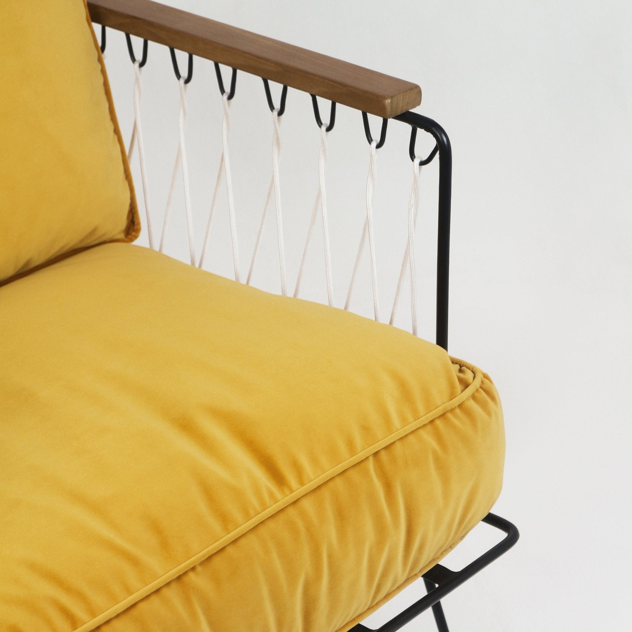 Close-up of a modern lounge chair showing a mustard yellow velvet seat cushion with piped edge, teak wood armrest, black metal frame and white rope woven back in a diamond pattern.