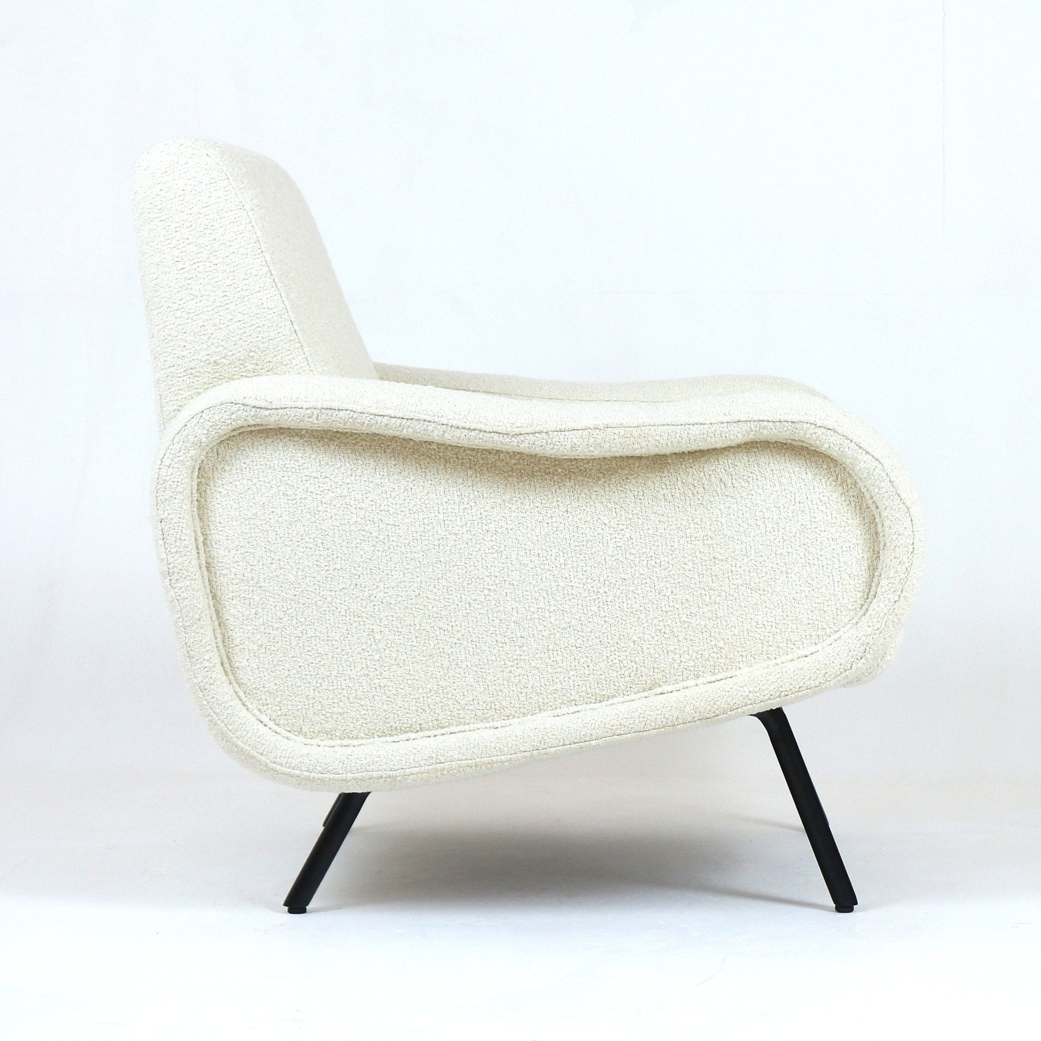 Side profile of a modern white boucle upholstered armchair with a curved low back, rounded padded arms and slender black metal legs against a plain white studio background.