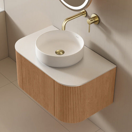 Floating fluted teak bathroom vanity with matte white stone top, round white ceramic vessel sink, and wall-mounted brushed brass faucet and pop-up drain.