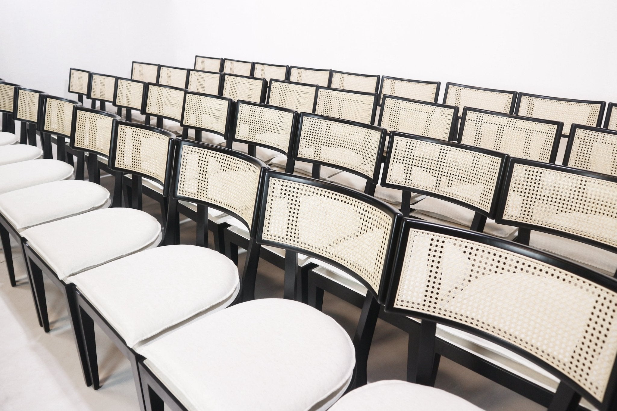 Multiple black-framed dining chairs arranged in rows, each with a woven natural rattan cane backrest and a cream upholstered seat, photographed from an angled perspective against a white background.