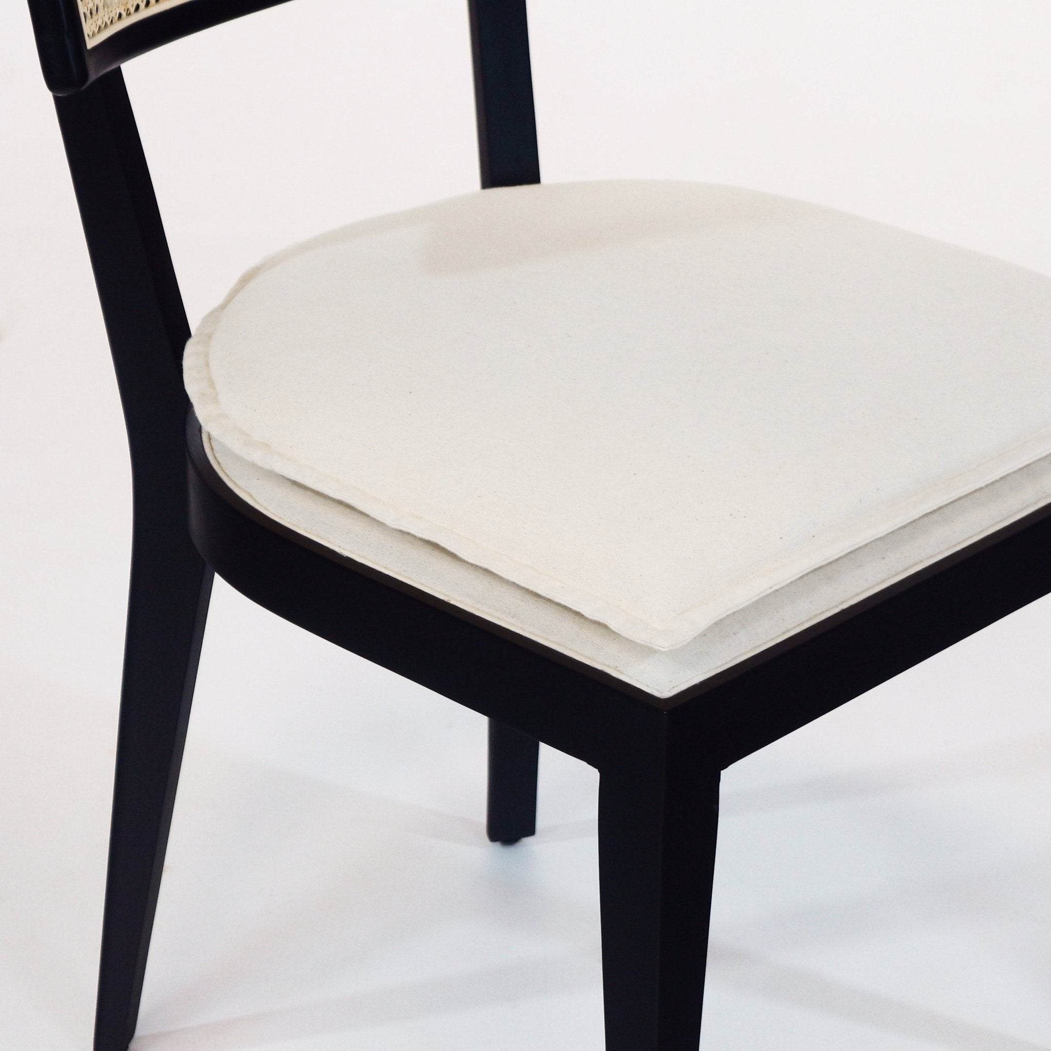 Close-up of a black solid-wood dining chair showing a cream fabric upholstered seat with a layered cushion and a visible woven rattan backrest detail.
