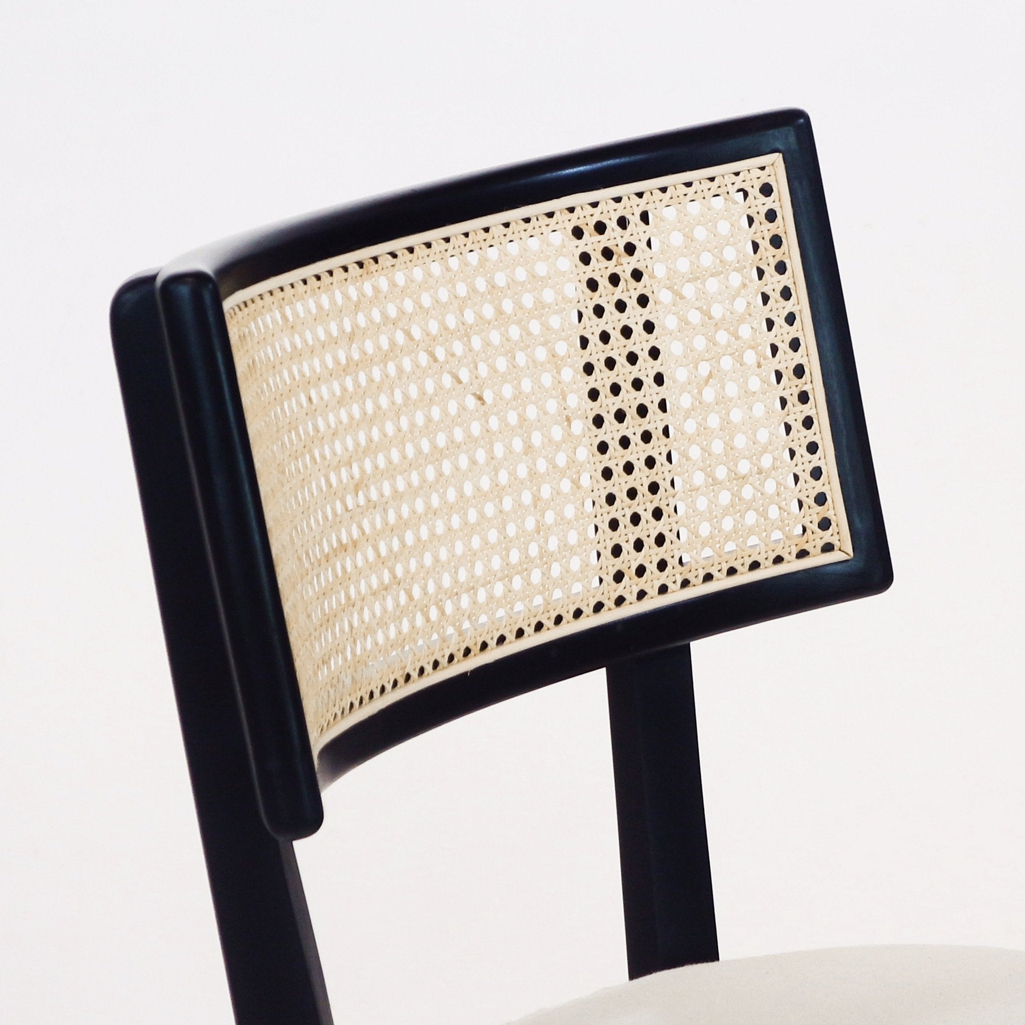 Close-up of a dining chair’s curved black solid-wood frame with a natural woven rattan (cane) backrest and a light upholstered seat visible at the bottom.