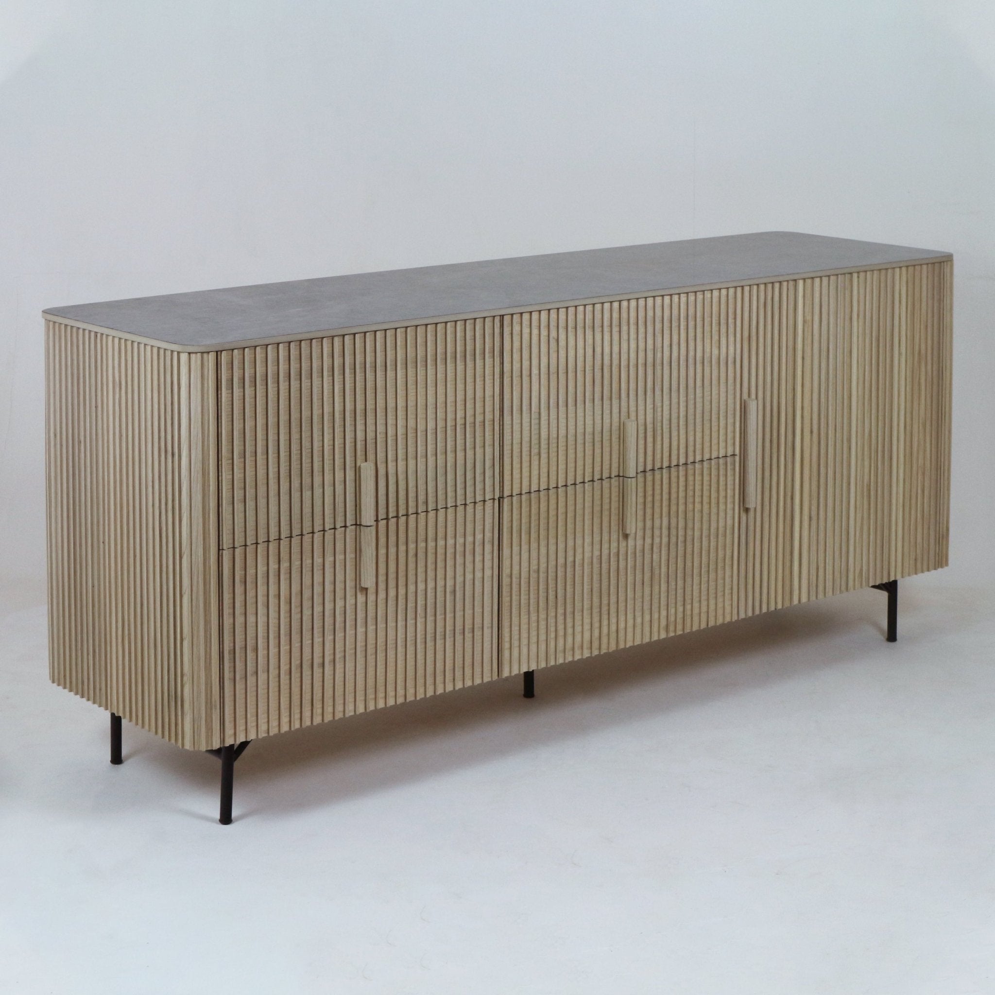 Long teak sideboard with vertical fluted wood doors, integrated vertical wooden handles, rounded corners, a gray stone-look top and slim black metal legs.
