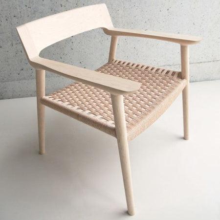 Light natural solid wood chair with a curved open back, flat armrests and a woven rope seat in a Scandinavian modern style, photographed on a neutral floor against a concrete wall.