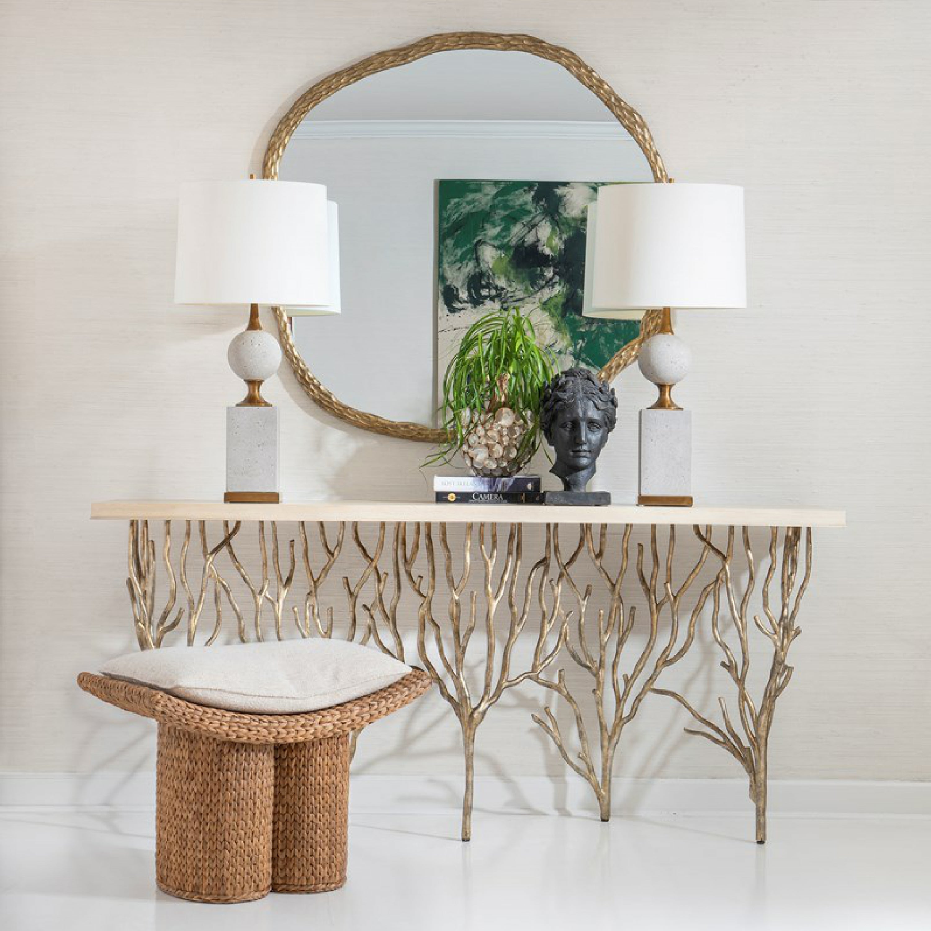 Natural handwoven hyacinth stool with a beige cushioned top sits in front of a modern-rustic gold-branch console table, round braided gold mirror and two white-shade lamps.