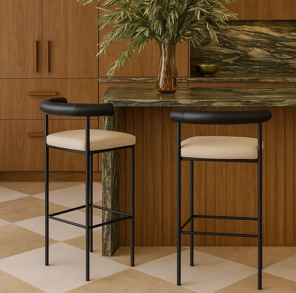 Two modern bar stools with matte black steel frames, curved black faux-leather low backs and light beige upholstered seats, positioned at a wood-paneled kitchen island with a green marble countertop and amber vase.
