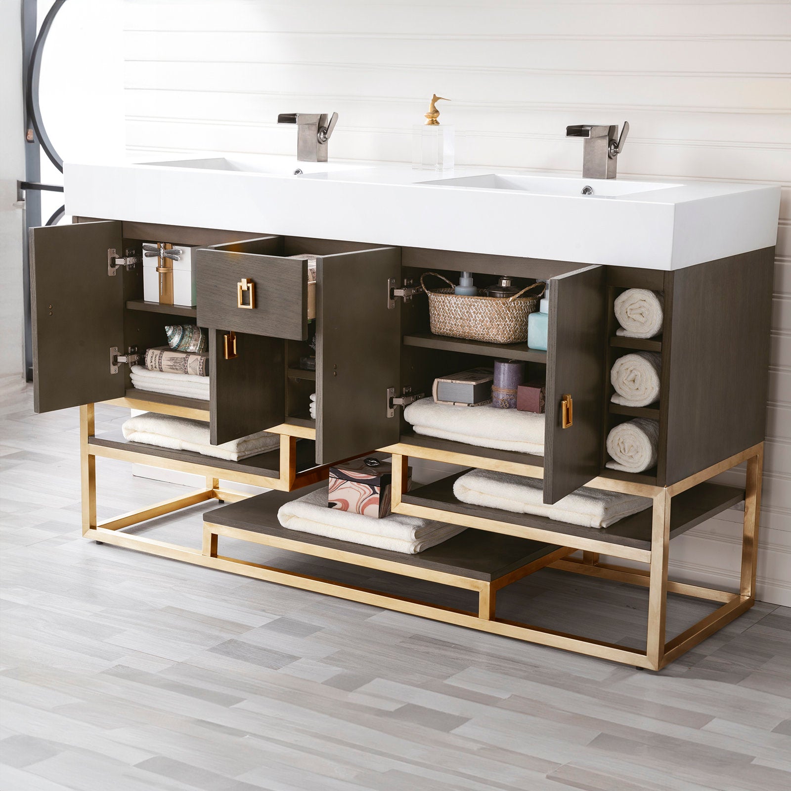 Modern solid-wood bathroom vanity with a white glossy stone composite double-sink top, dark wood cabinet and gold-finish metal base, open shelves and cabinets staged with rolled towels, basket and two metal faucets.