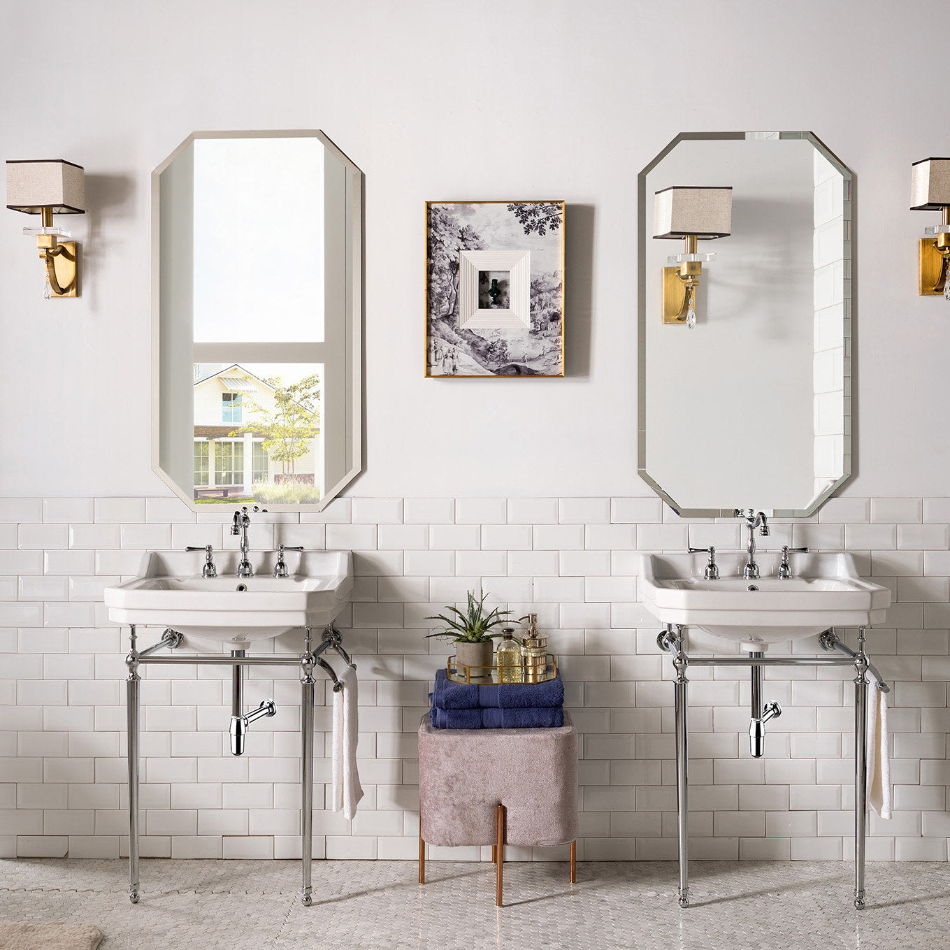 Pair of freestanding white ceramic console sinks with chrome metal legs and exposed plumbing against a stone-colored wall and white subway tile, flanked by octagonal mirrors, brass wall sconces and a small stool with folded towels.